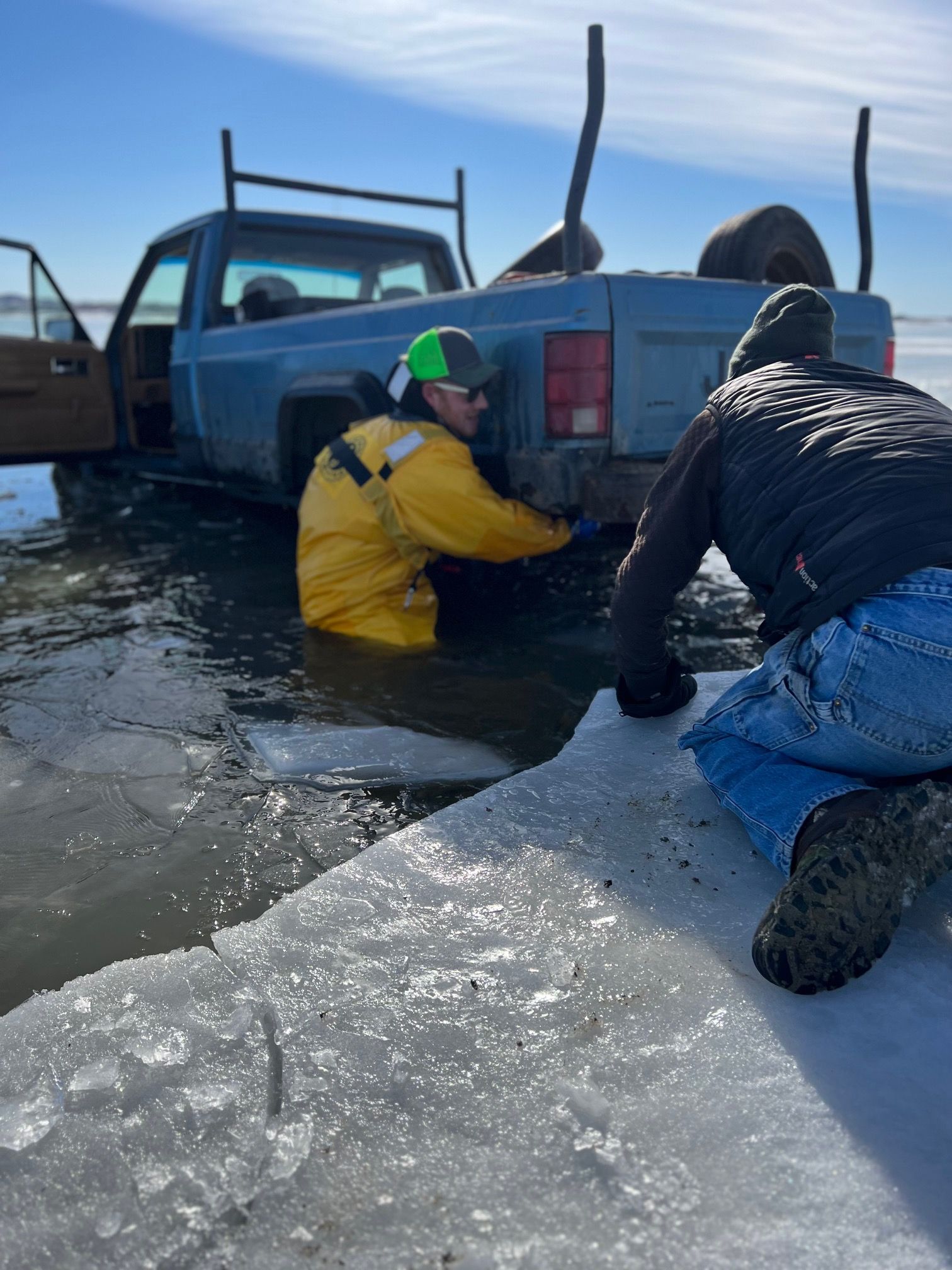 Two people try to free a blue pickup truck stuck in icy water on a sunny day.