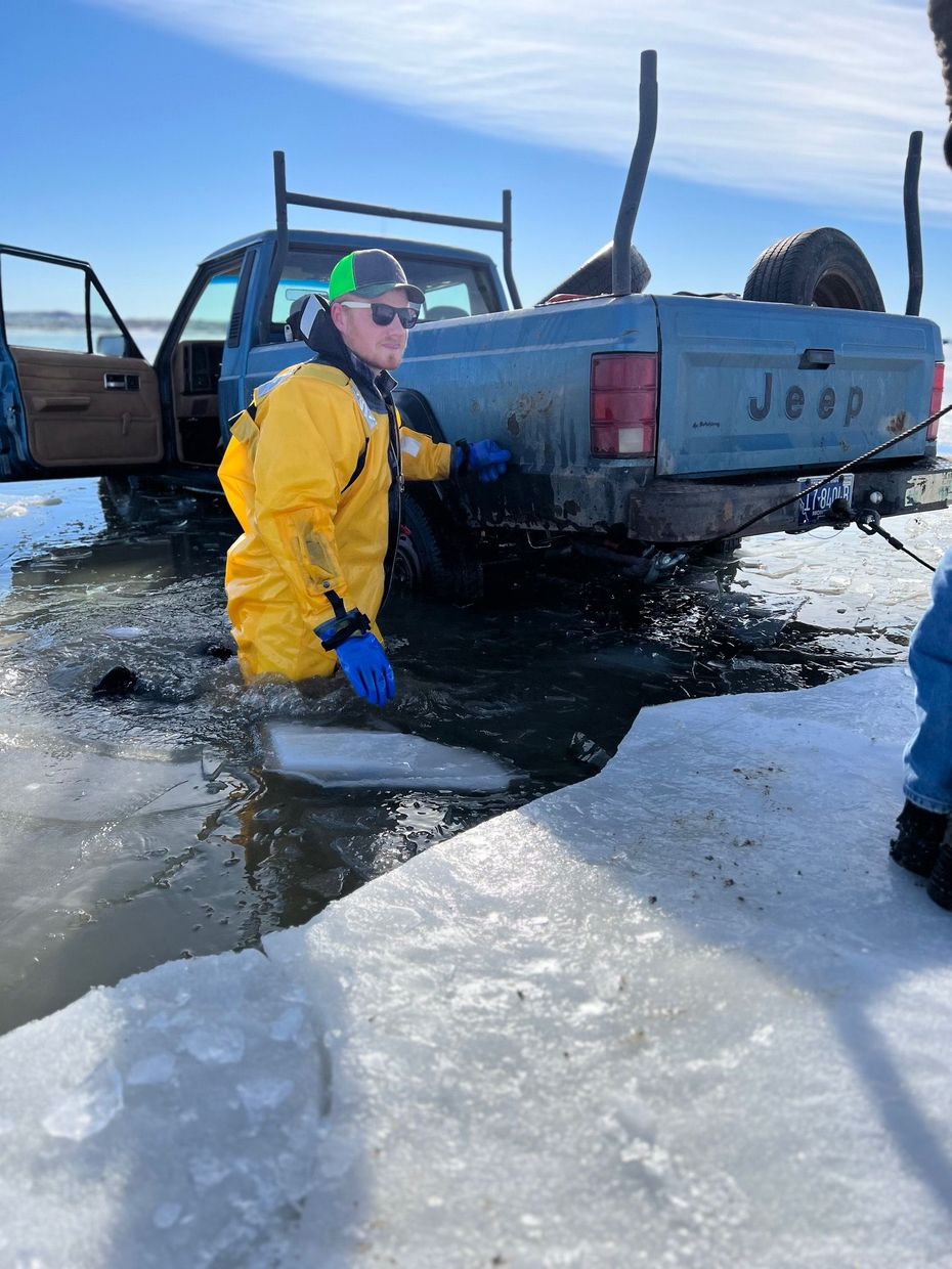 Man in yellow suit standing in icy water near a Jeep truck stuck in ice, sunny day.