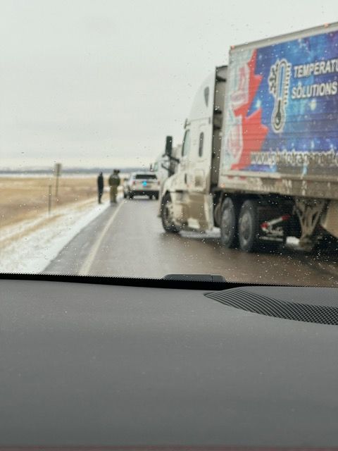 Semi-truck and vehicles stopped on a snowy road; two people and a police car are visible.