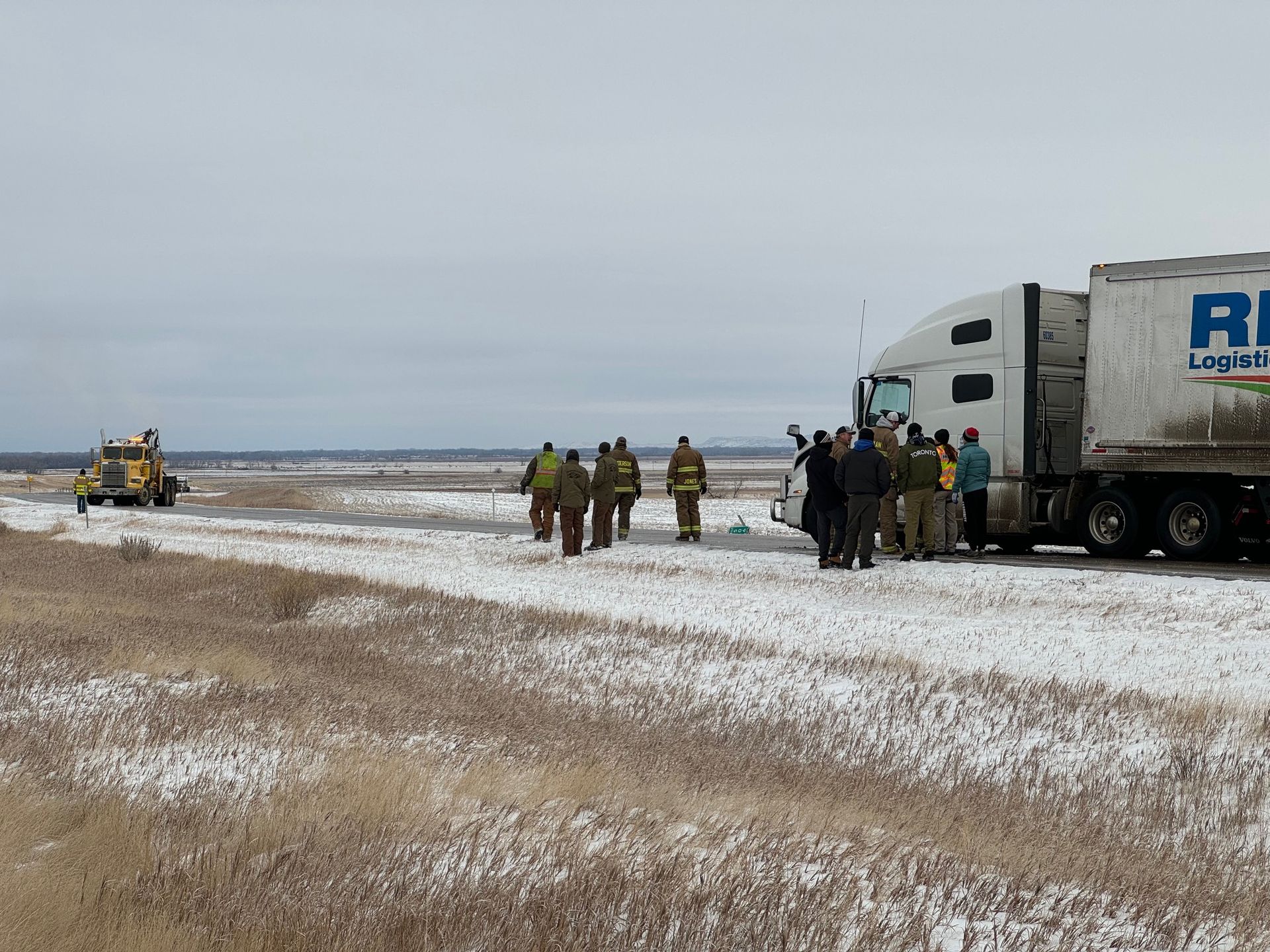 Group of people near semi-truck in snowy field; wrecker in distance.