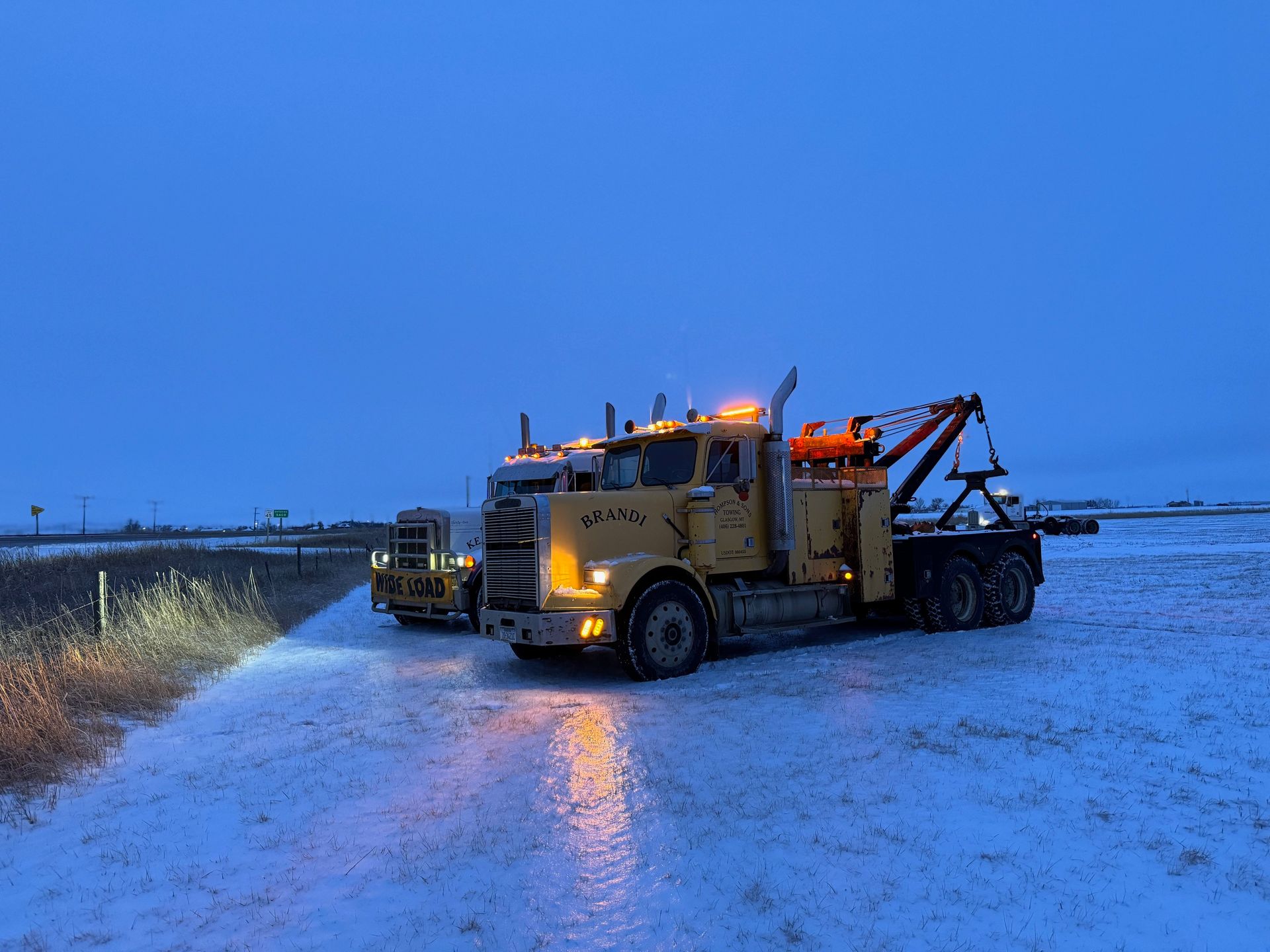 Two yellow tow trucks parked on a snow-covered road under a dusky sky, lights on.