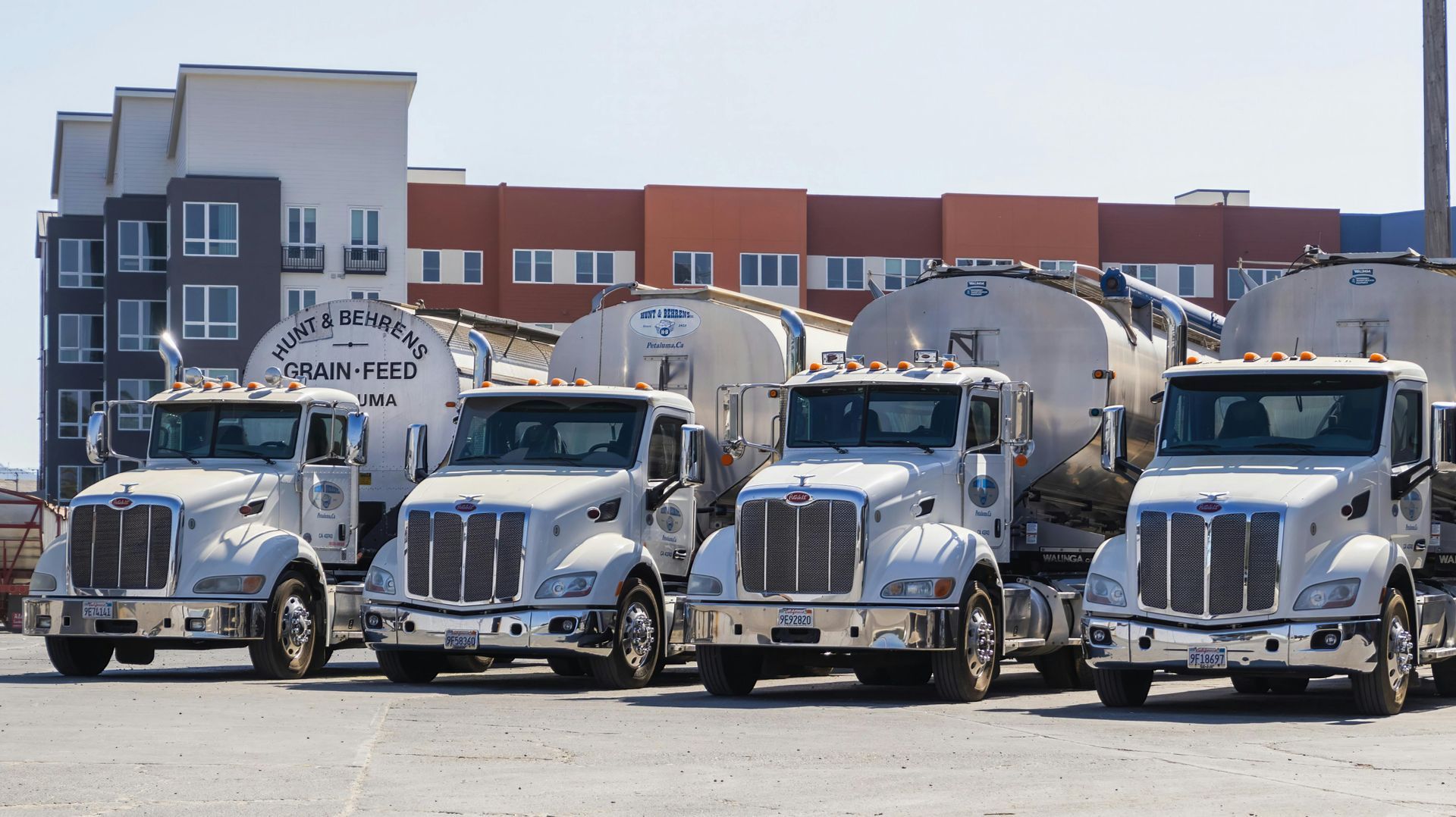 Four silver tanker trucks parked in a row in front of an industrial building