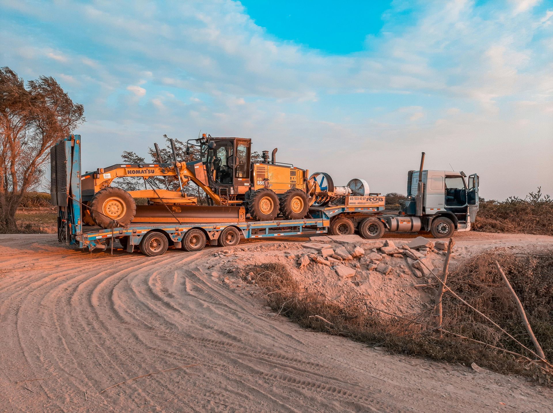 Yellow excavator and dump truck on a lowboy trailer along a dirt road under a cloudy sky