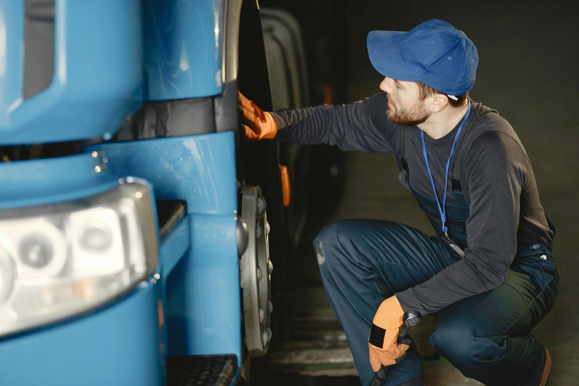 A person in a blue cap, dark uniform, and orange gloves crouches to inspect the side of a large blue truck.