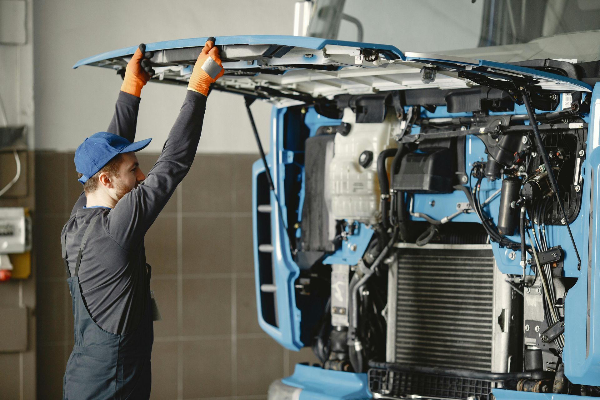 A mechanic in a cap and uniform lifts the hood of a large blue truck in a repair shop.