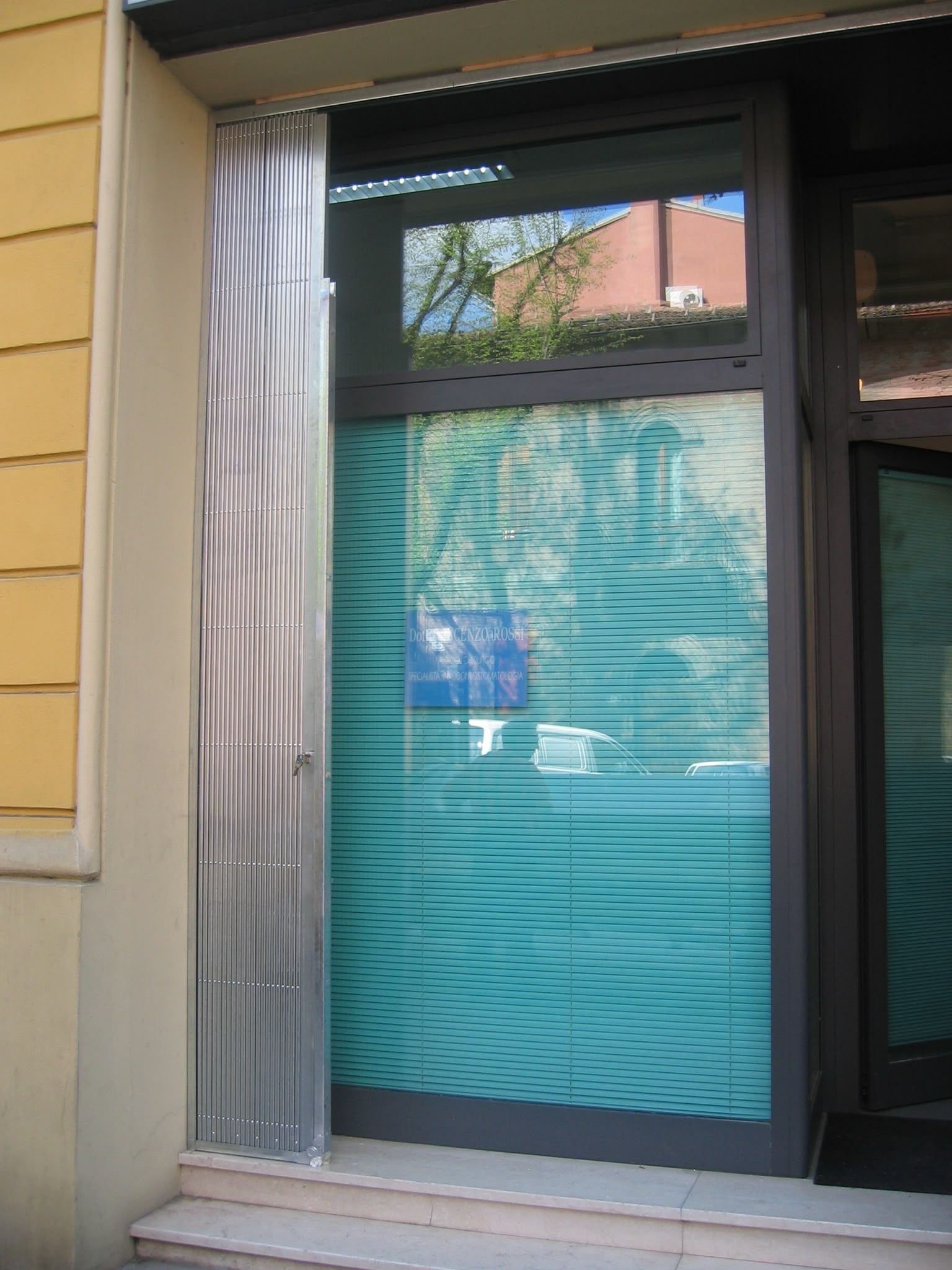 Vetrina con persiane blu e tenda bianca, incorniciata da un rivestimento beige.