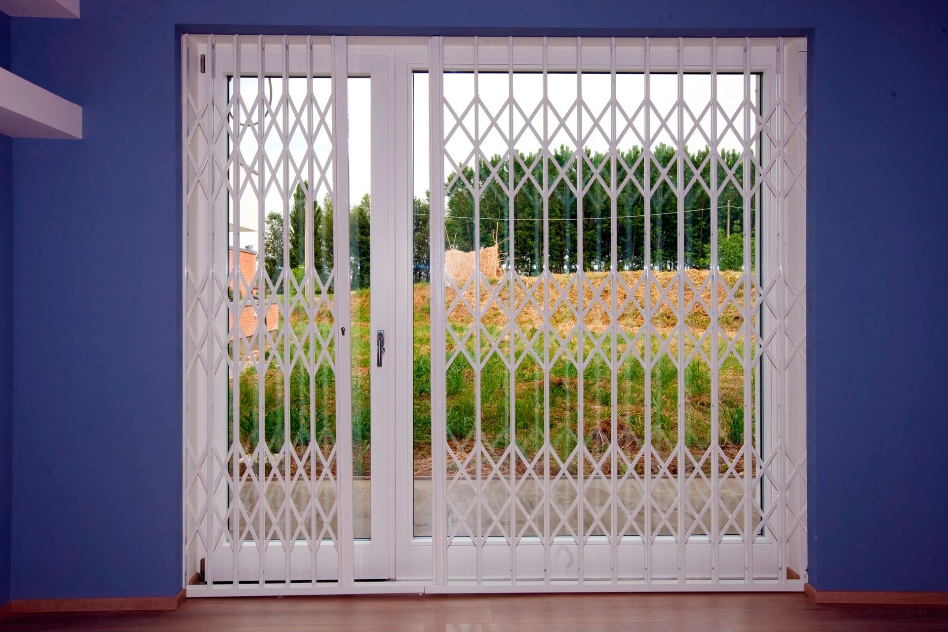Porte pieghevoli bianche con grate di sicurezza a griglia che si aprono su un giardino fiorito e alberi all'esterno.