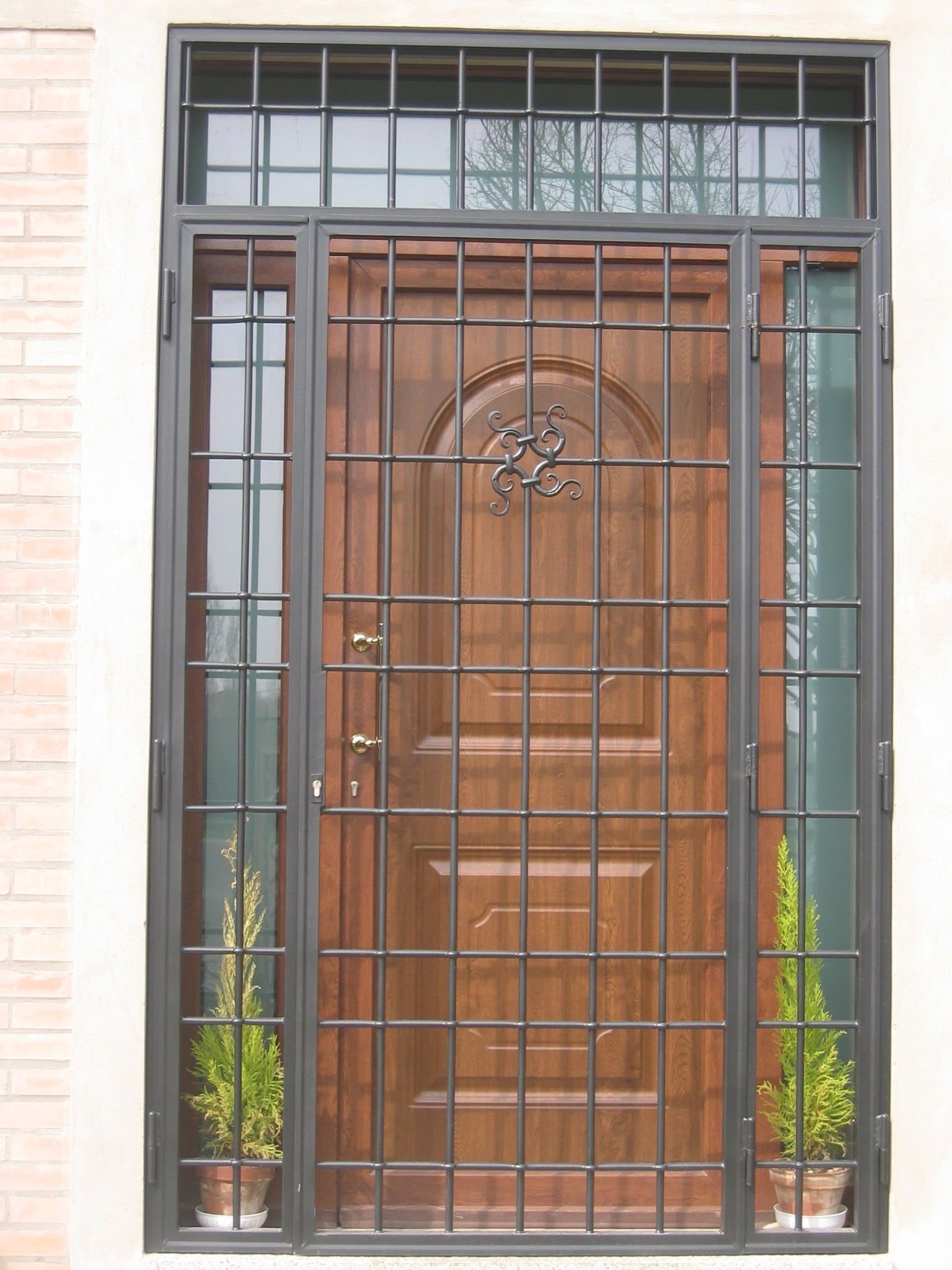 Porta d'ingresso in legno protetta da sbarre di sicurezza in metallo nero, con pannelli laterali satinati e piccole piante in vaso su entrambi i lati.