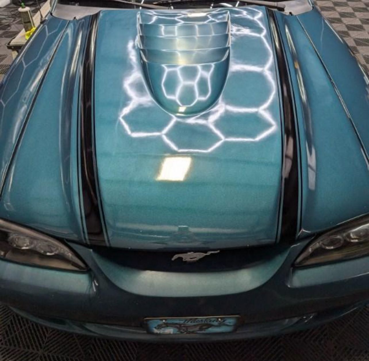 Teal car hood with a scoop and black racing stripes reflecting honeycomb patterns.