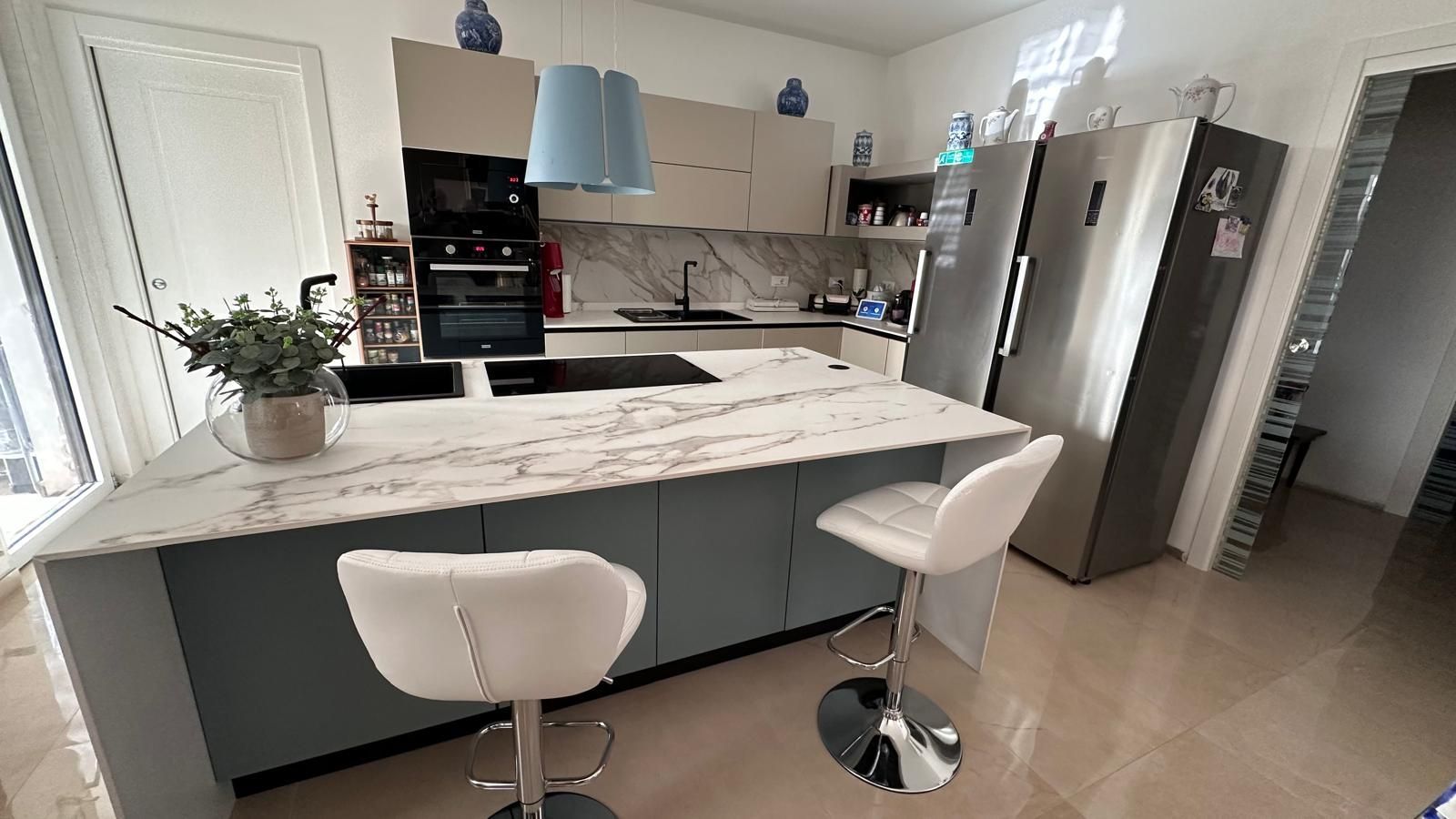 Cucina moderna con isola, piano di lavoro in marmo, elettrodomestici in acciaio inossidabile e due sgabelli da bar bianchi.