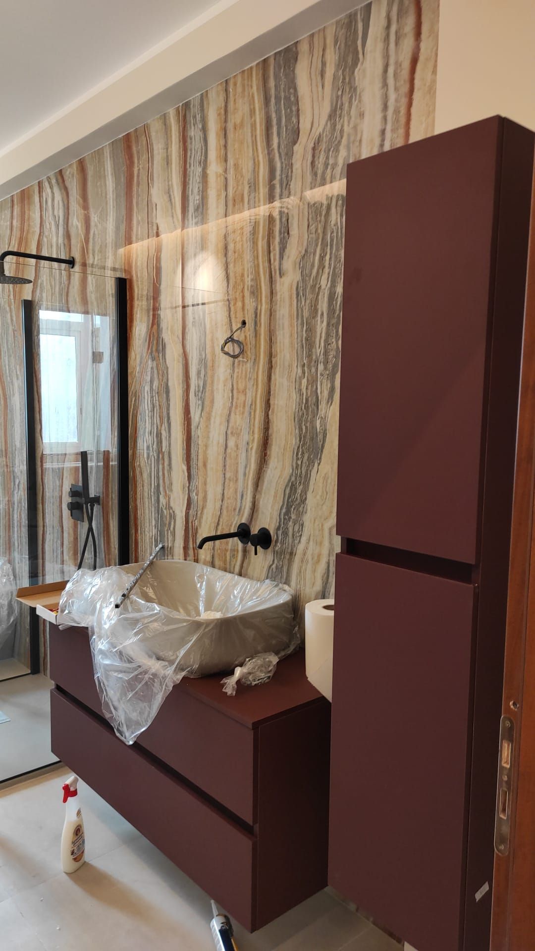 Bagno con piastrelle effetto marmo, mobiletto e mobiletto alto color marrone, sanitari neri e lavandino moderno.