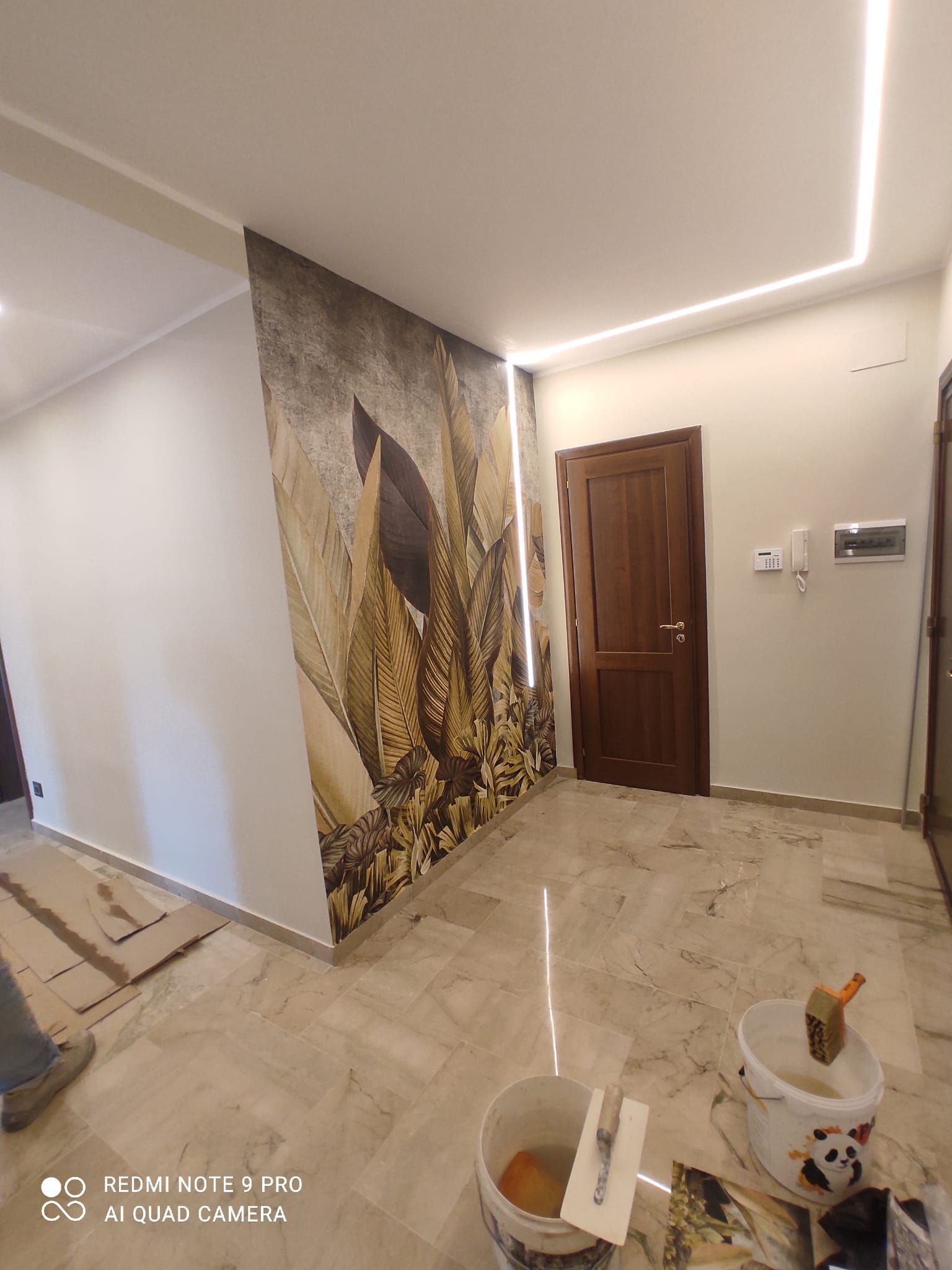 Un corridoio interno con pavimenti in marmo, una parete a mosaico, una porta marrone e secchi con gli attrezzi.