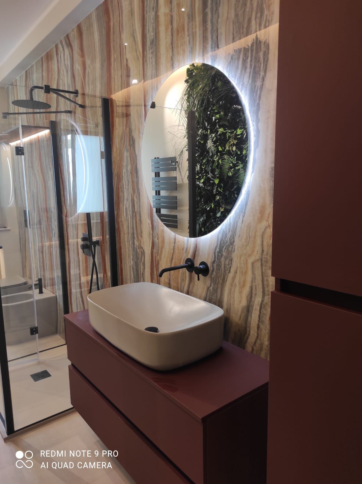 Bagno moderno con lavabo a appoggio, specchio rotondo con verde e mobiletto rossastro.
