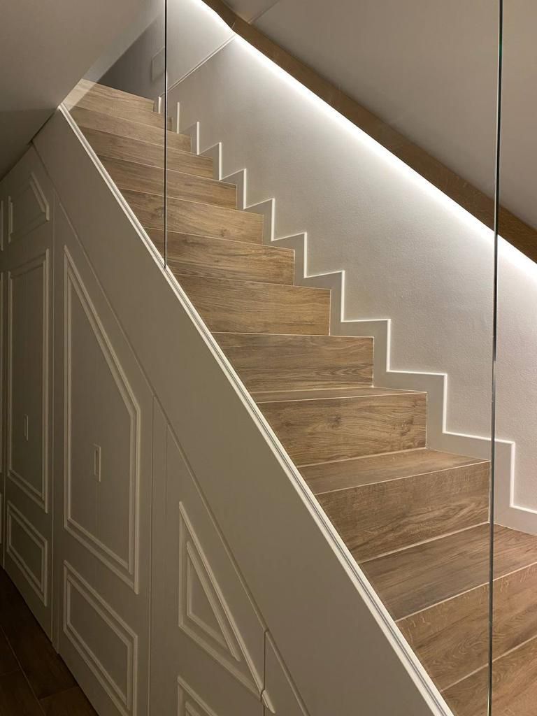 Scala con gradini in legno e illuminazione a LED. Parete bianca con pannelli a sinistra, ringhiera in vetro a destra.