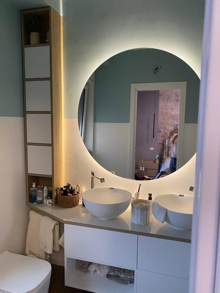Bagno con pareti bianche e verde acqua, specchio rotondo, doppi lavandini e un mobiletto alto.
