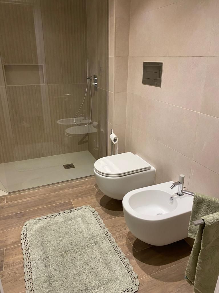 Bagno moderno con doccia, WC e bidet. Pareti piastrellate beige, pavimento effetto legno, tappeto verde e asciugamano.