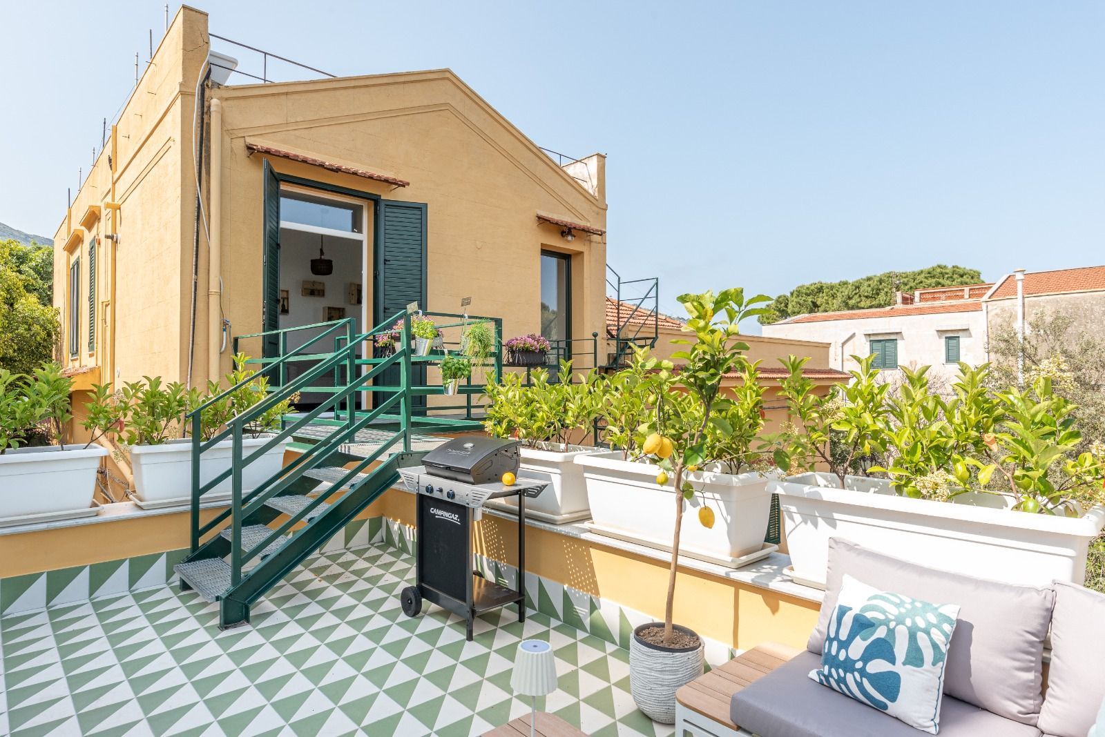 Edificio giallo con scala verde che conduce a un patio sul tetto. Dispone di barbecue, alberi di limone e divano.