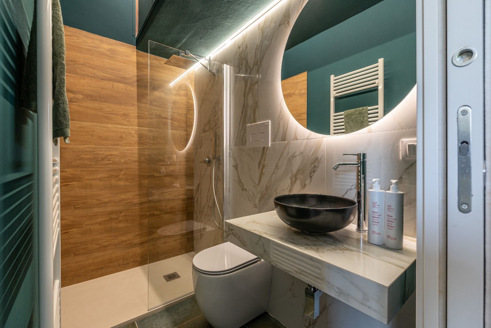 Bagno con doccia effetto legno, mobile lavabo sospeso, lavandino nero, grande specchio illuminato, pareti color verde acqua.
