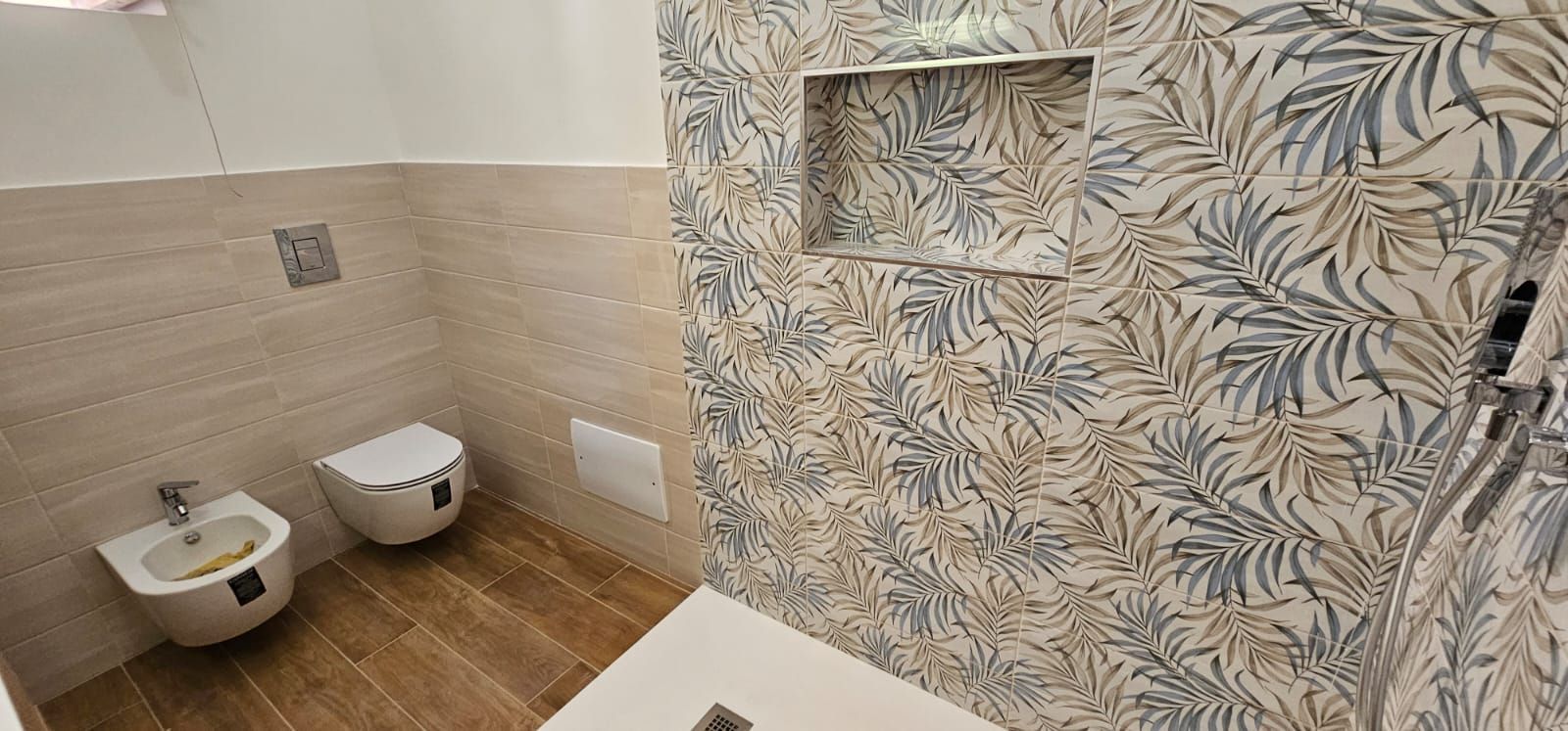 Bagno con carta da parati floreale, WC, bidet, pavimento effetto legno e mensola incassata.