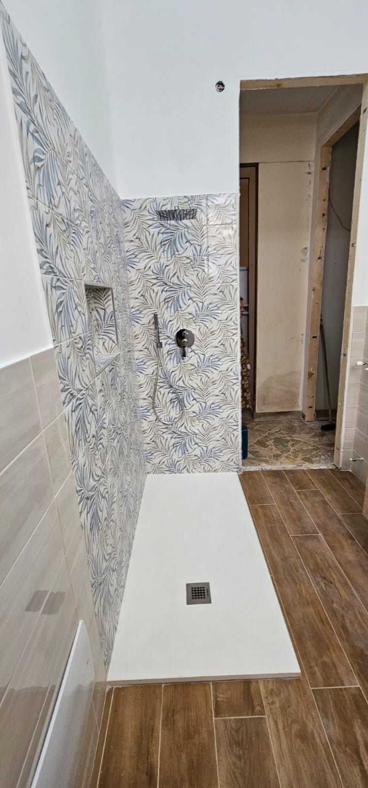 Doccia con parete piastrellata blu e pavimento effetto legno. Piatto doccia bianco e sanitari neri. Porta aperta.