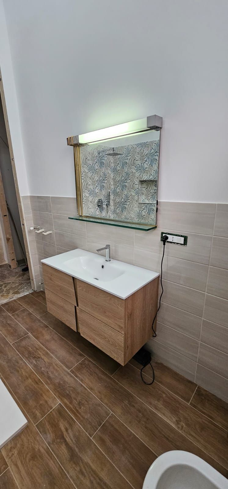 Bagno con mobile lavabo in legno, specchio e WC. Pavimento in piastrelle marroni e pareti beige chiaro.