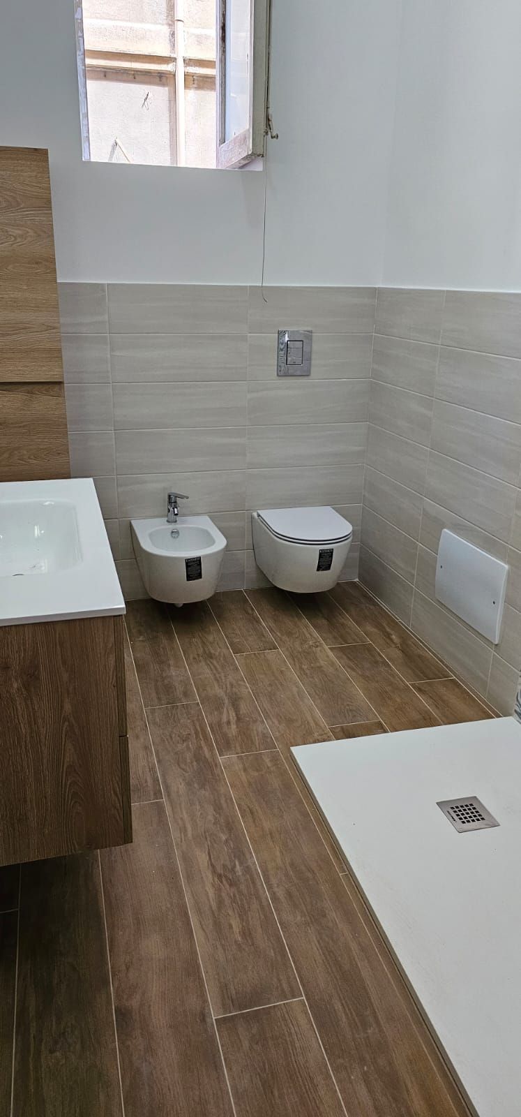 Bagno con lavandino e WC. Pavimento effetto legno. Piastrelle bianco sporco. WC e bidet bianchi.