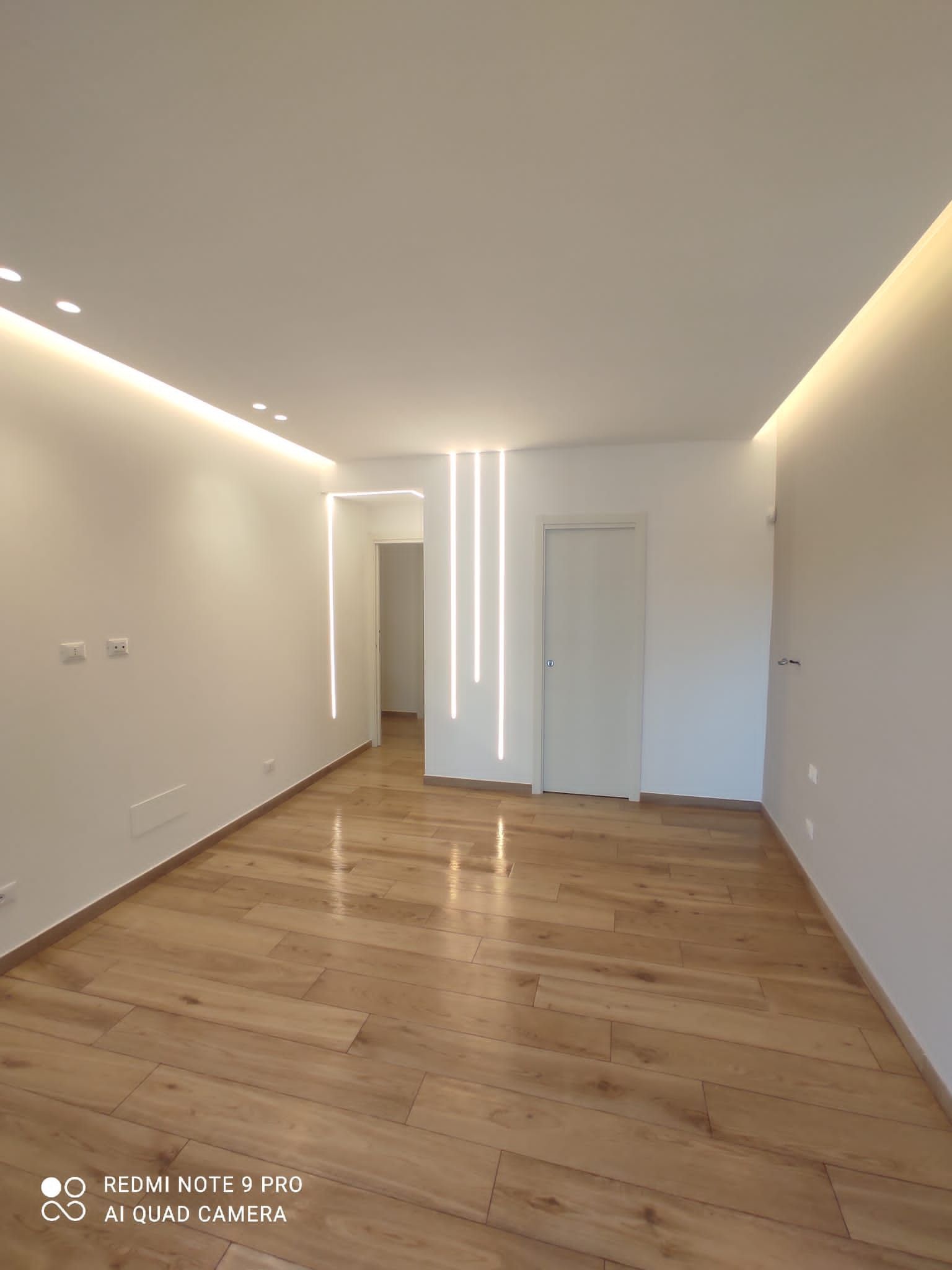 Stanza vuota con pavimenti in legno e pareti bianche. Illuminazione a incasso. Porte e corridoio a vista.