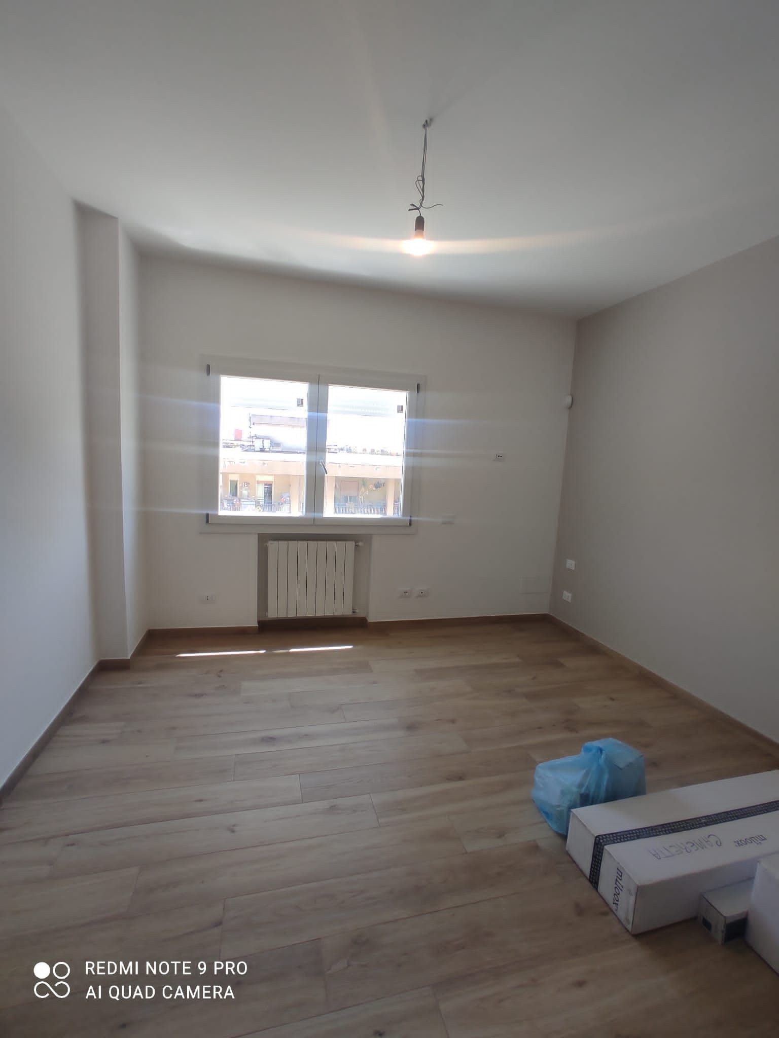 Stanza vuota con pavimenti in legno, una finestra e fili scoperti che pendono dal soffitto.