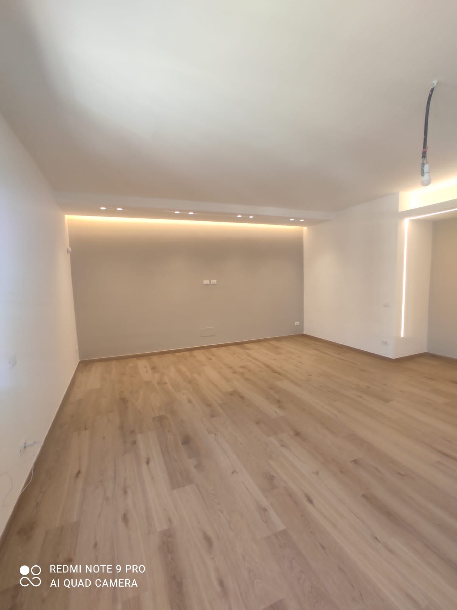 Stanza vuota con pavimenti in legno chiaro, illuminazione da incasso e una parete decorativa grigia.