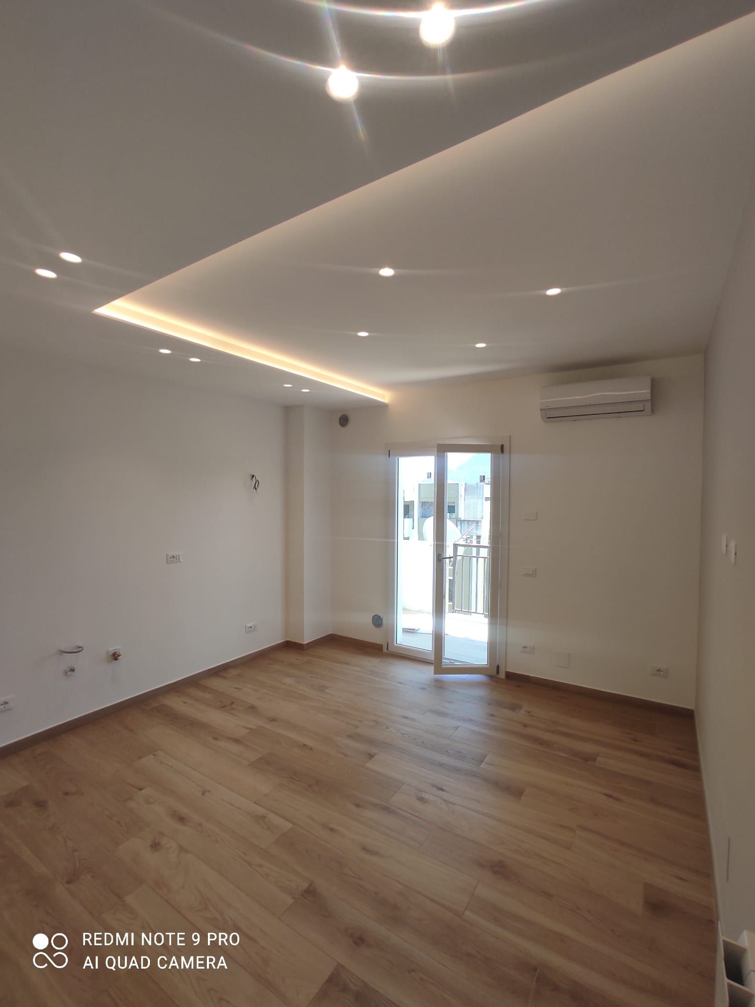 Stanza vuota con pavimenti in legno chiaro, pareti bianche, faretti da incasso nel soffitto e accesso al balcone.