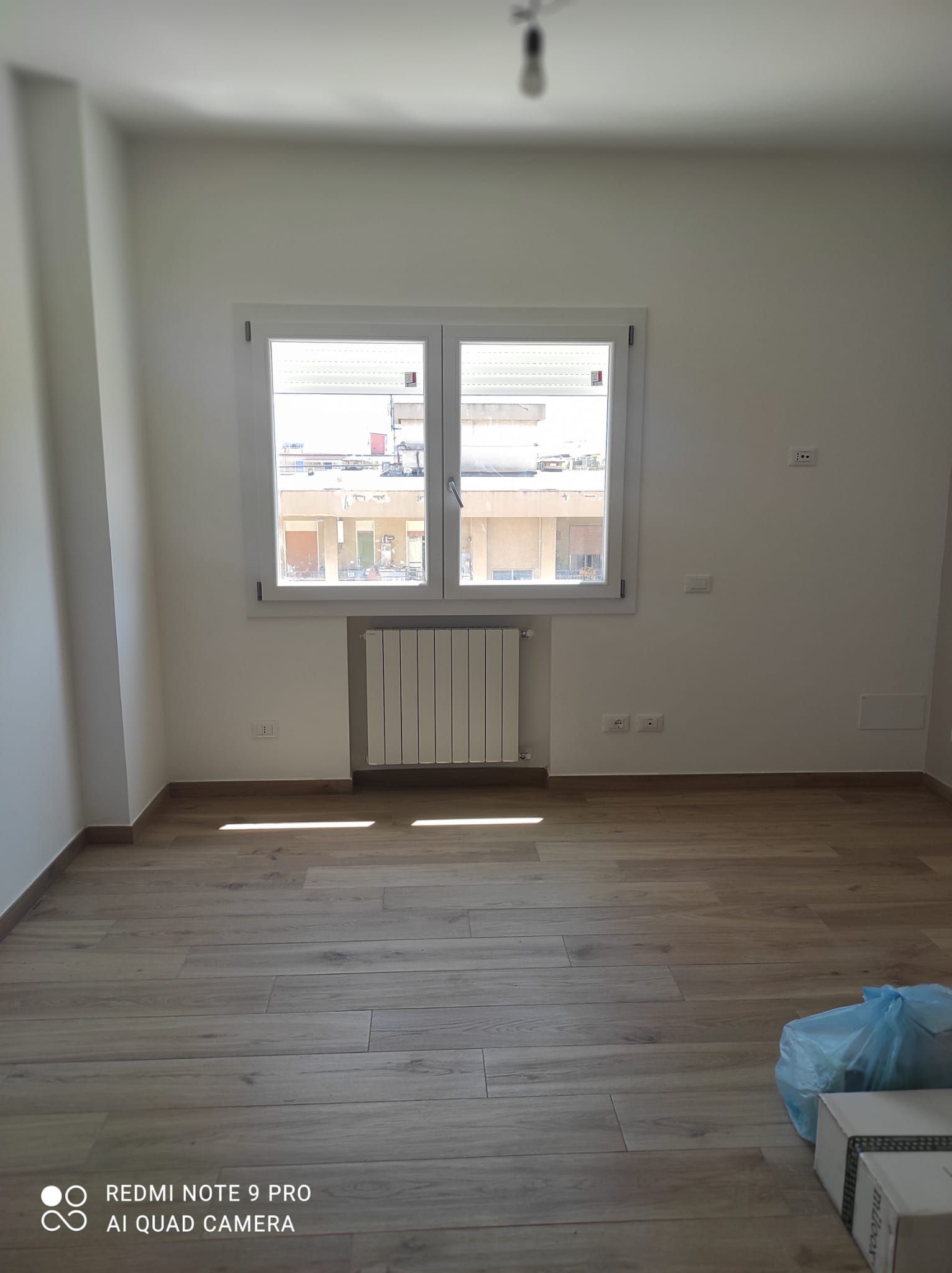 Stanza vuota con finestra, termosifone e pavimento in legno. Luce naturale e intensa.