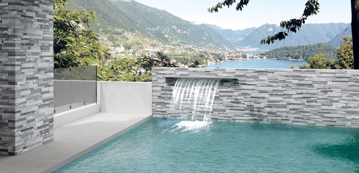 Piscina con cascata che si affaccia sul lago e sulle montagne. Pareti in pietra grigia e acqua verde brillante.