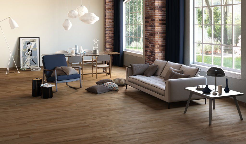 Soggiorno con pavimenti in legno, parete in mattoni e ampie finestre; divano, sedie e opere d'arte.