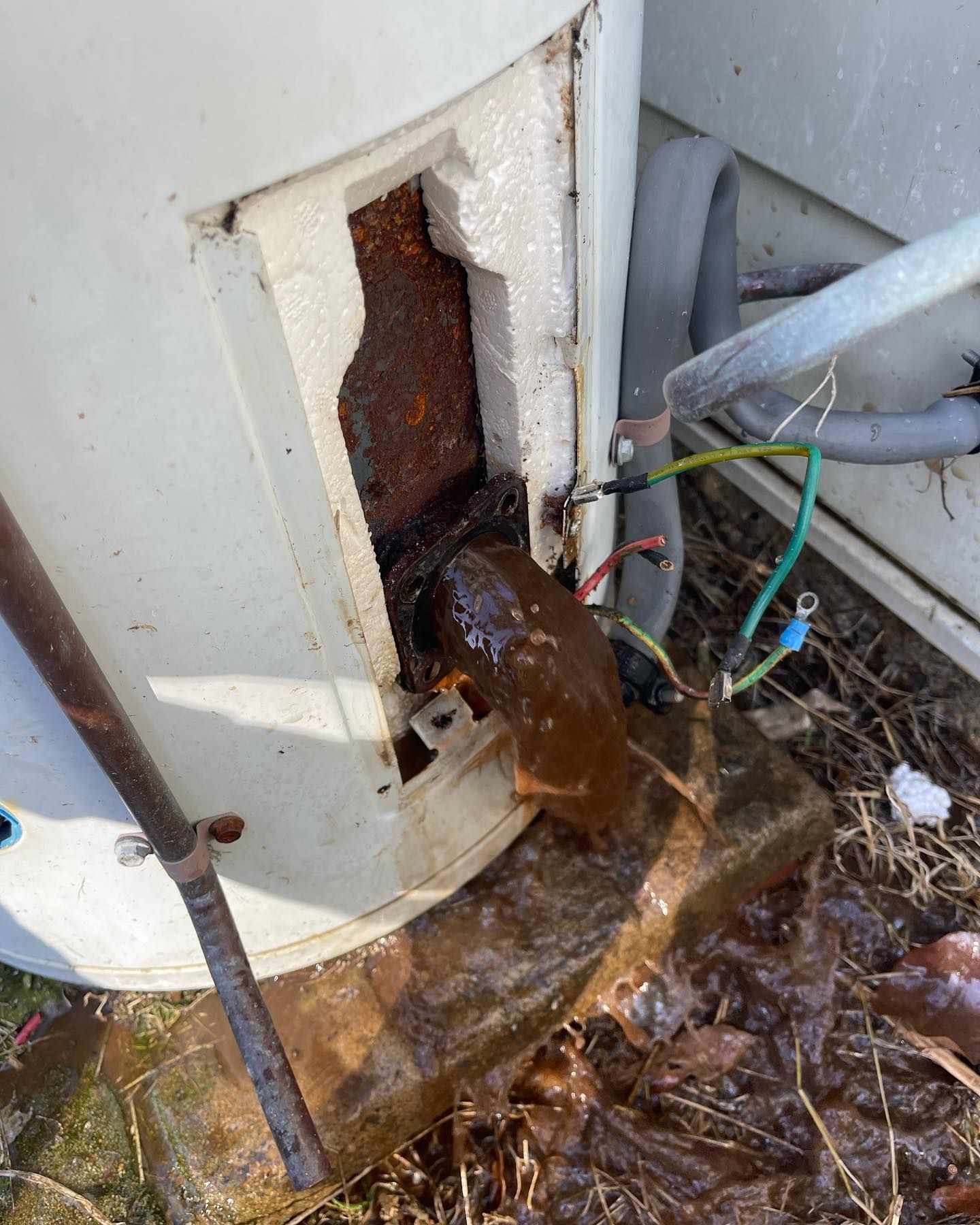 Rusty, damaged outdoor utility unit with exposed wiring and muddy water leaking onto the ground