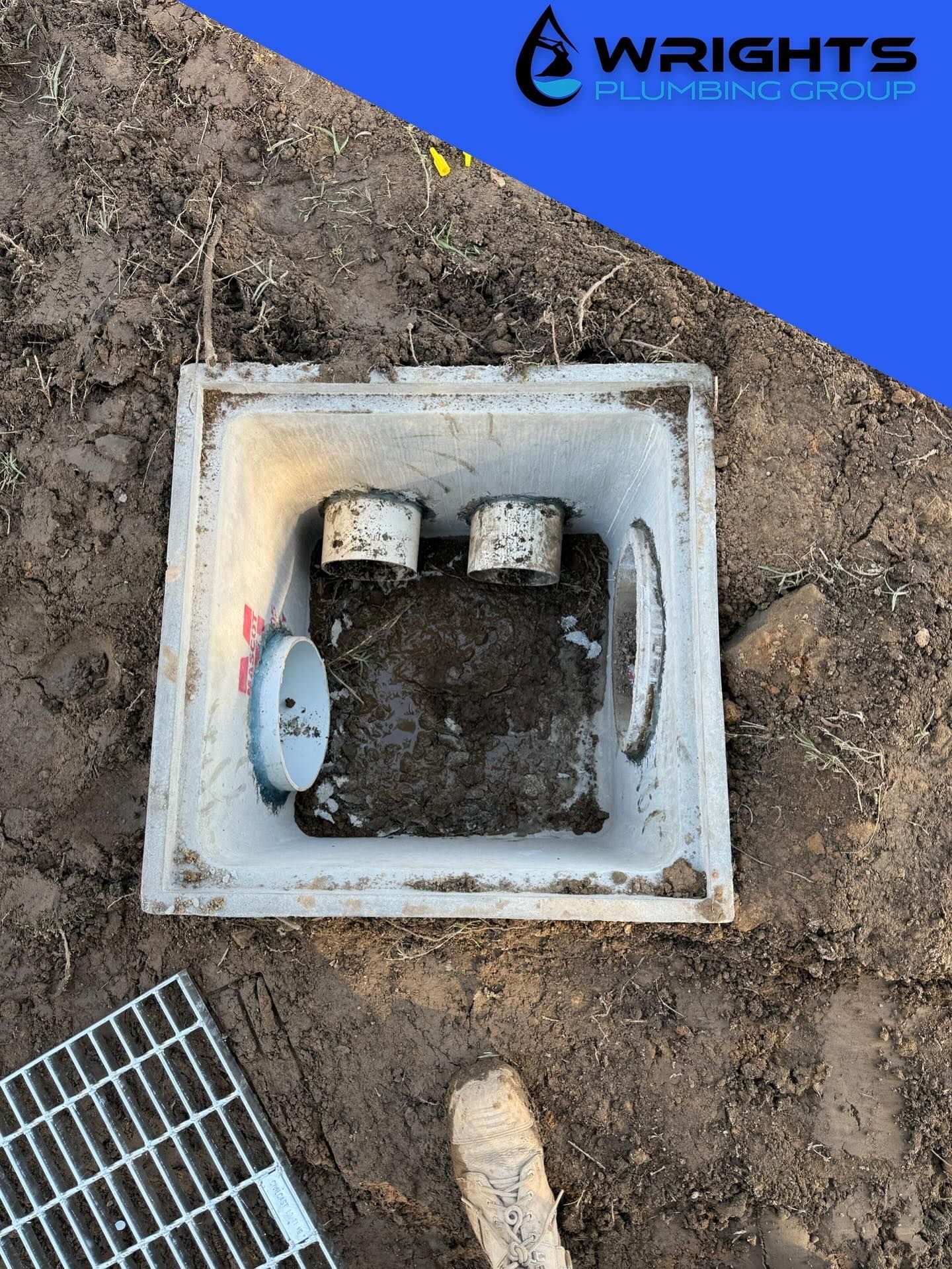 Open utility box with exposed pipes in dirt, next to a metal grate and branded blue corner watermark.