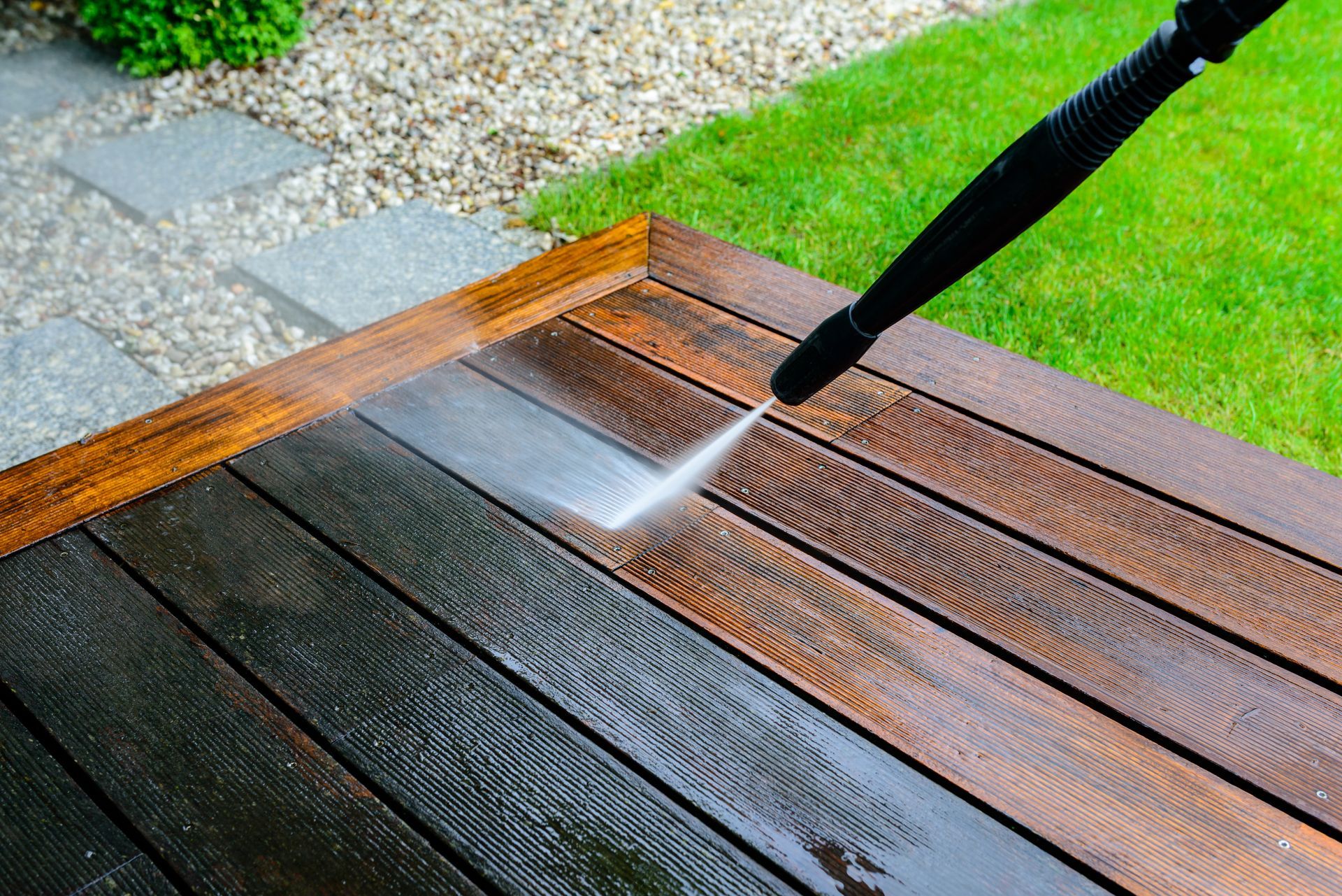 Pressure washing a wooden deck with a black spray wand, cleaning the wet boards near gravel and grass.
