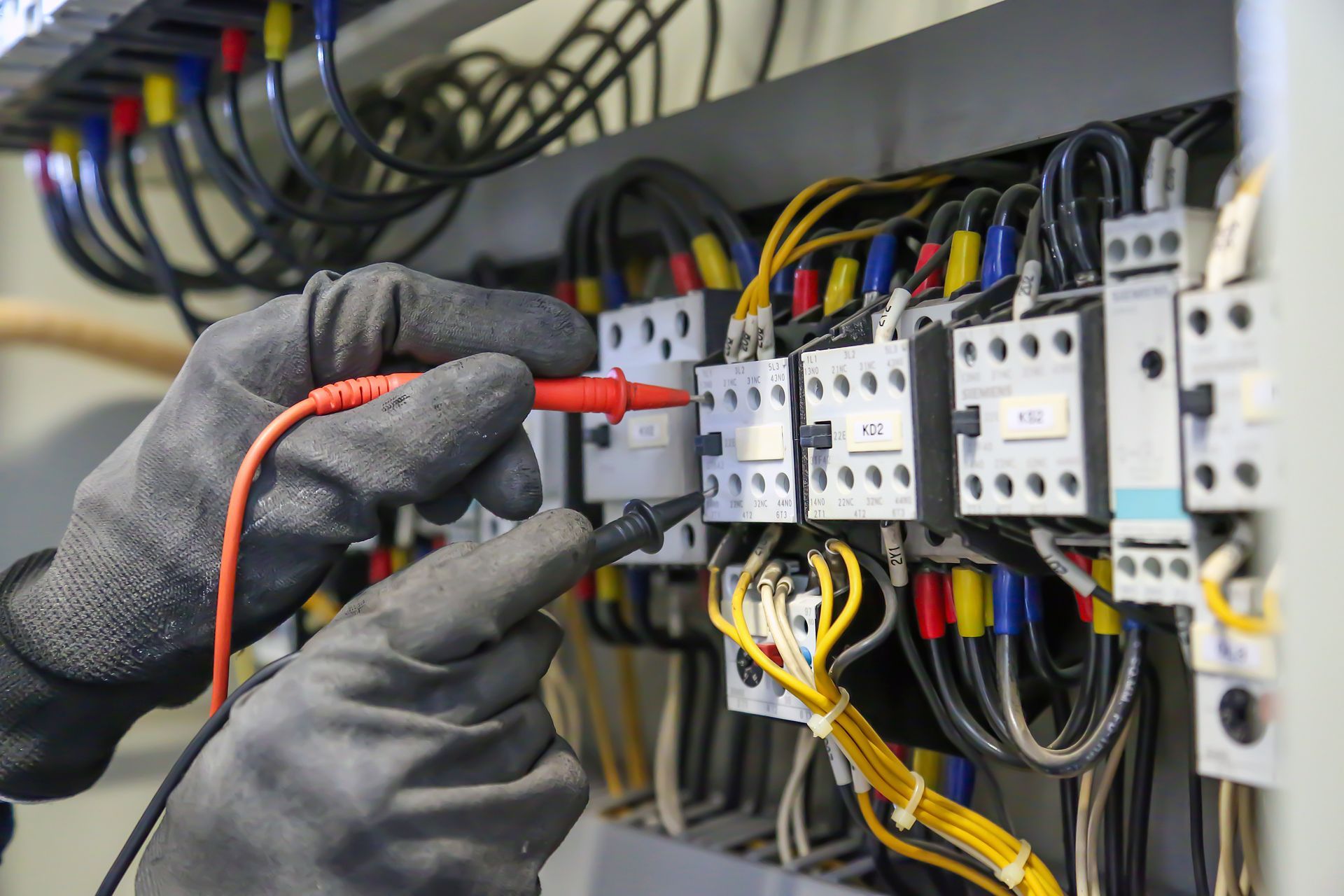 Gloved hands testing electrical wiring in a control panel with multicolored cables and circuit breakers
