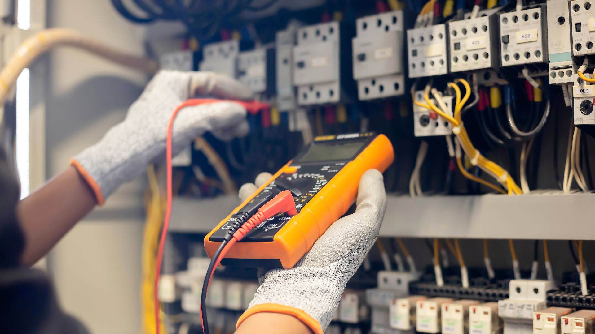 Gloved hands testing wiring in an electrical control panel with a multimeter