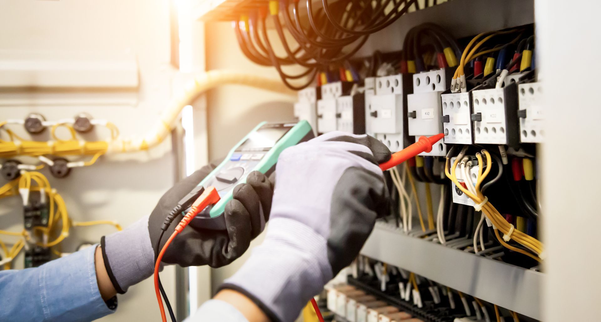 Gloved hands testing electrical wiring in an open control panel with a multimeter