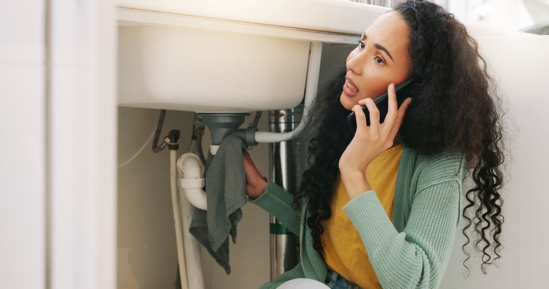 Woman with curly hair crouches under a sink, talking on a phone while inspecting plumbing.