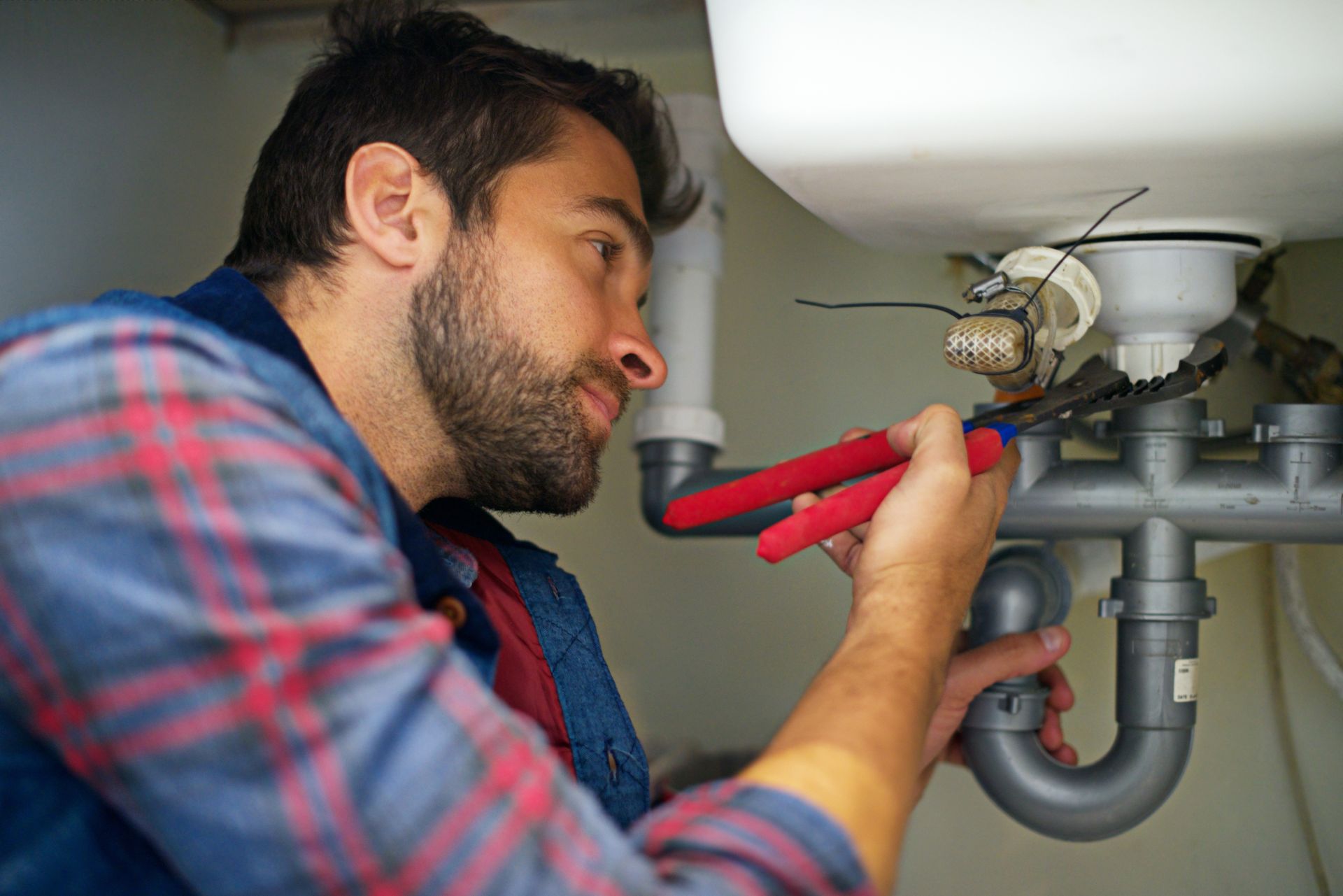 Plumber using red pliers to repair under-sink plumbing pipes and faucet