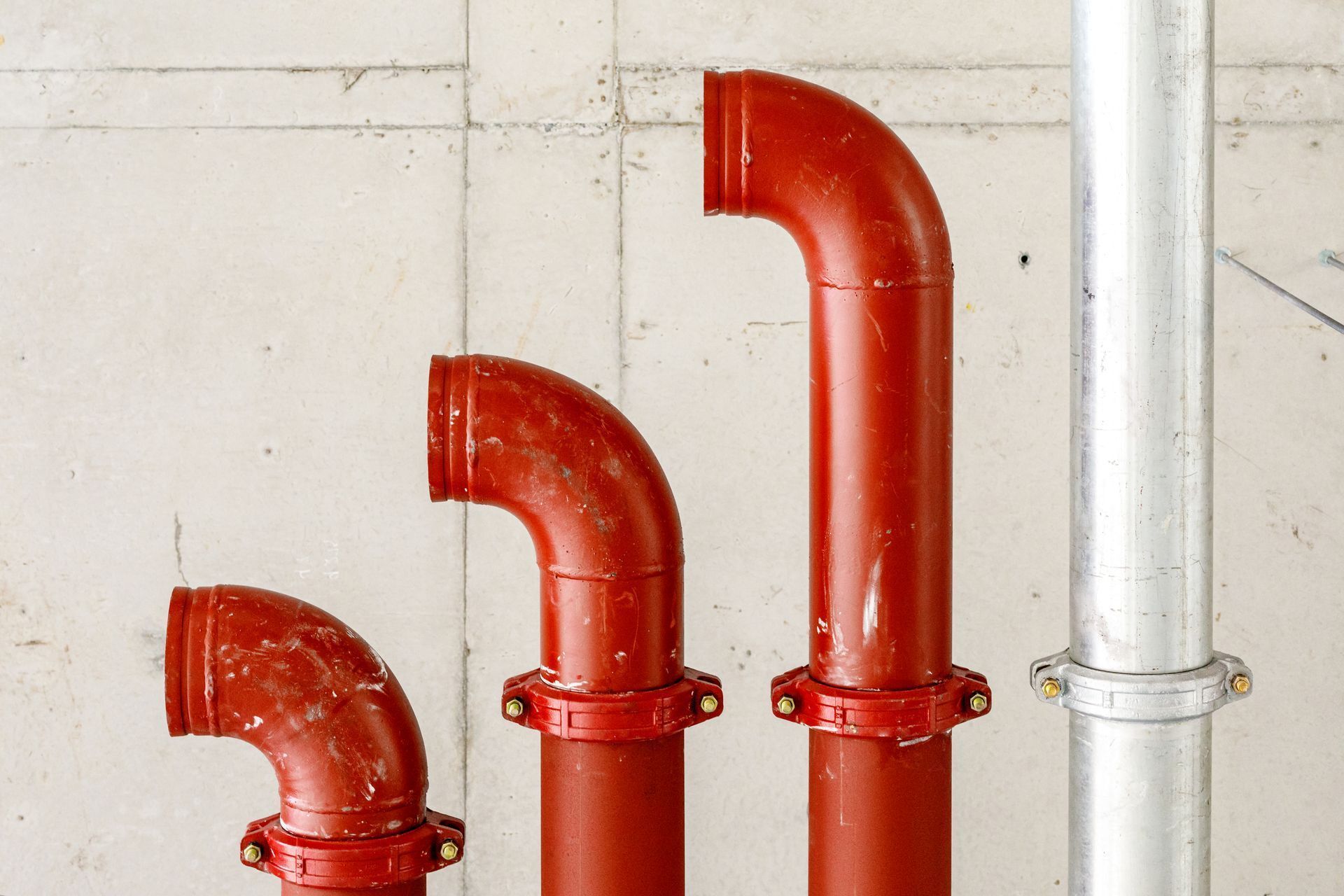 Three red industrial pipes of varying heights beside a silver pipe against a concrete wall