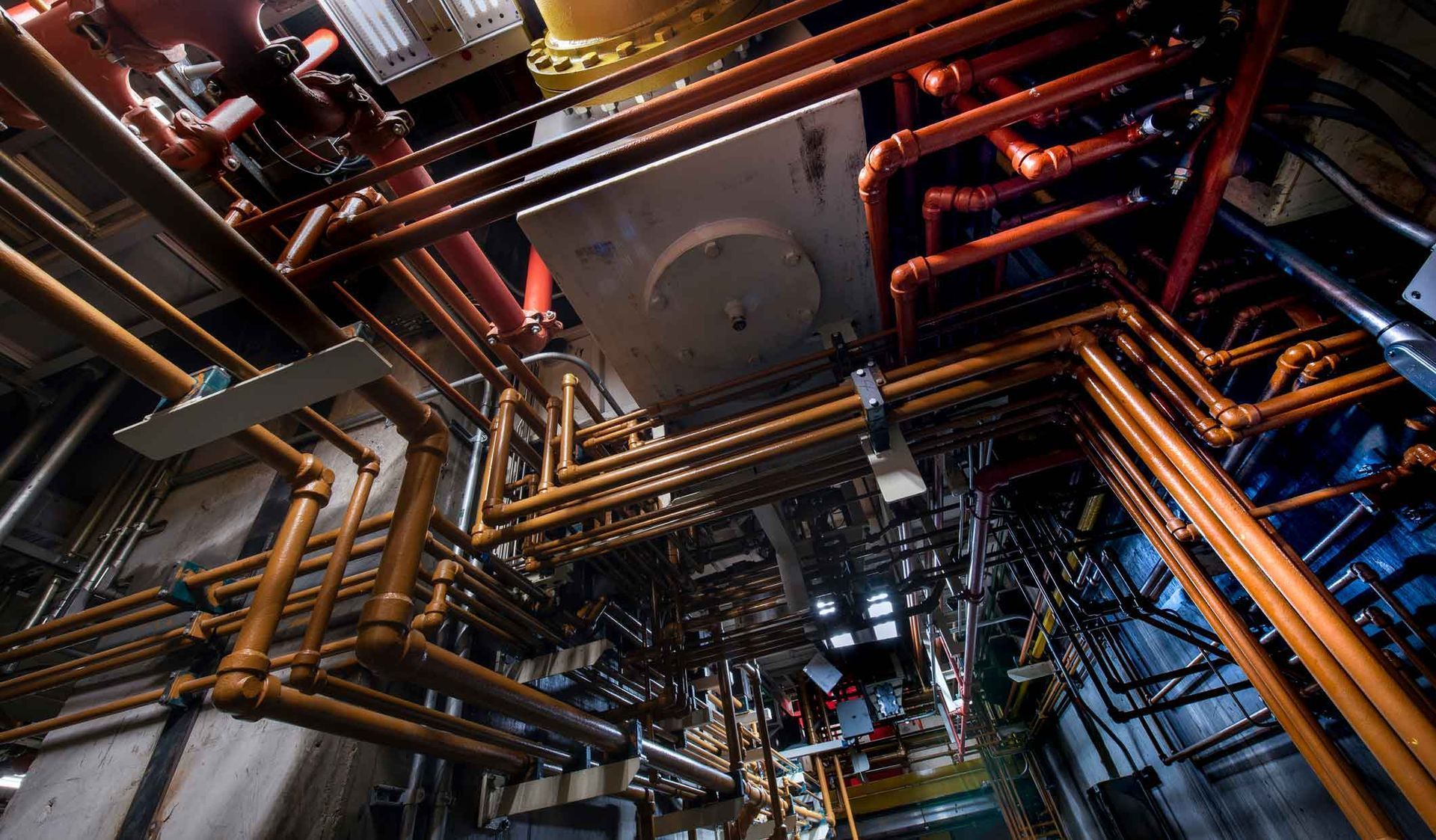Dense industrial pipe network with copper and orange pipes in a dark mechanical ceiling space