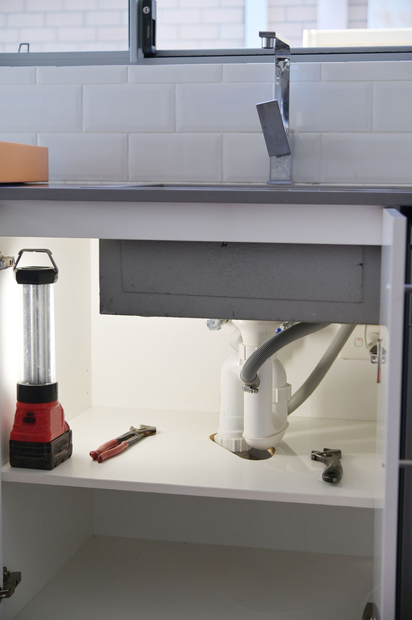 Under-sink cabinet under renovation with exposed plumbing and a work light on the left.