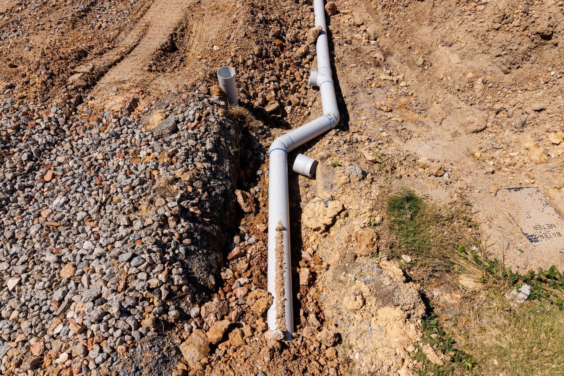 Damaged white drainage pipe running through a dirt trench beside a gravel pile