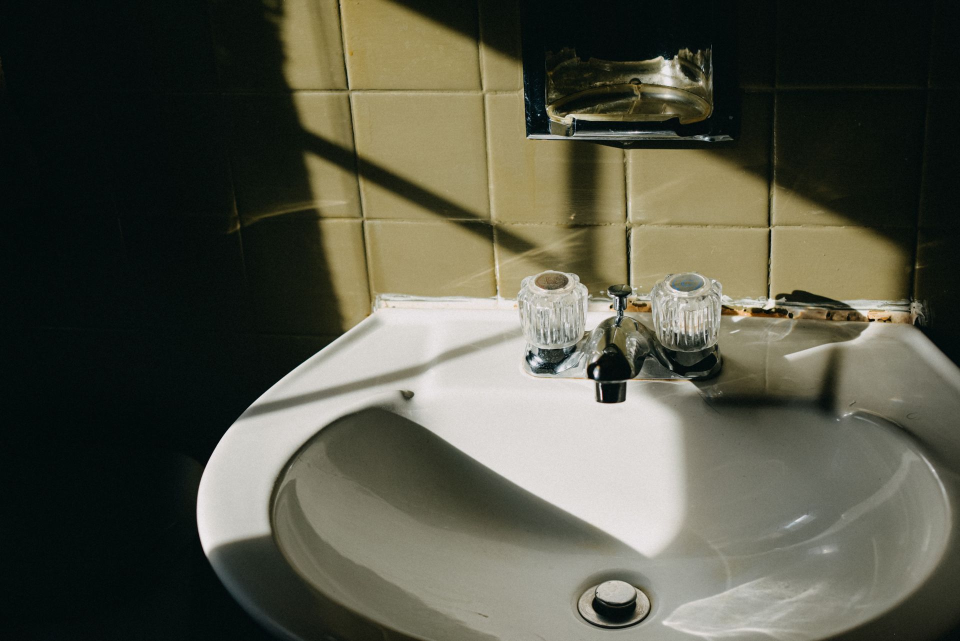 White bathroom sink with two faucets and a mirror above, lit by sunlight and shadow.