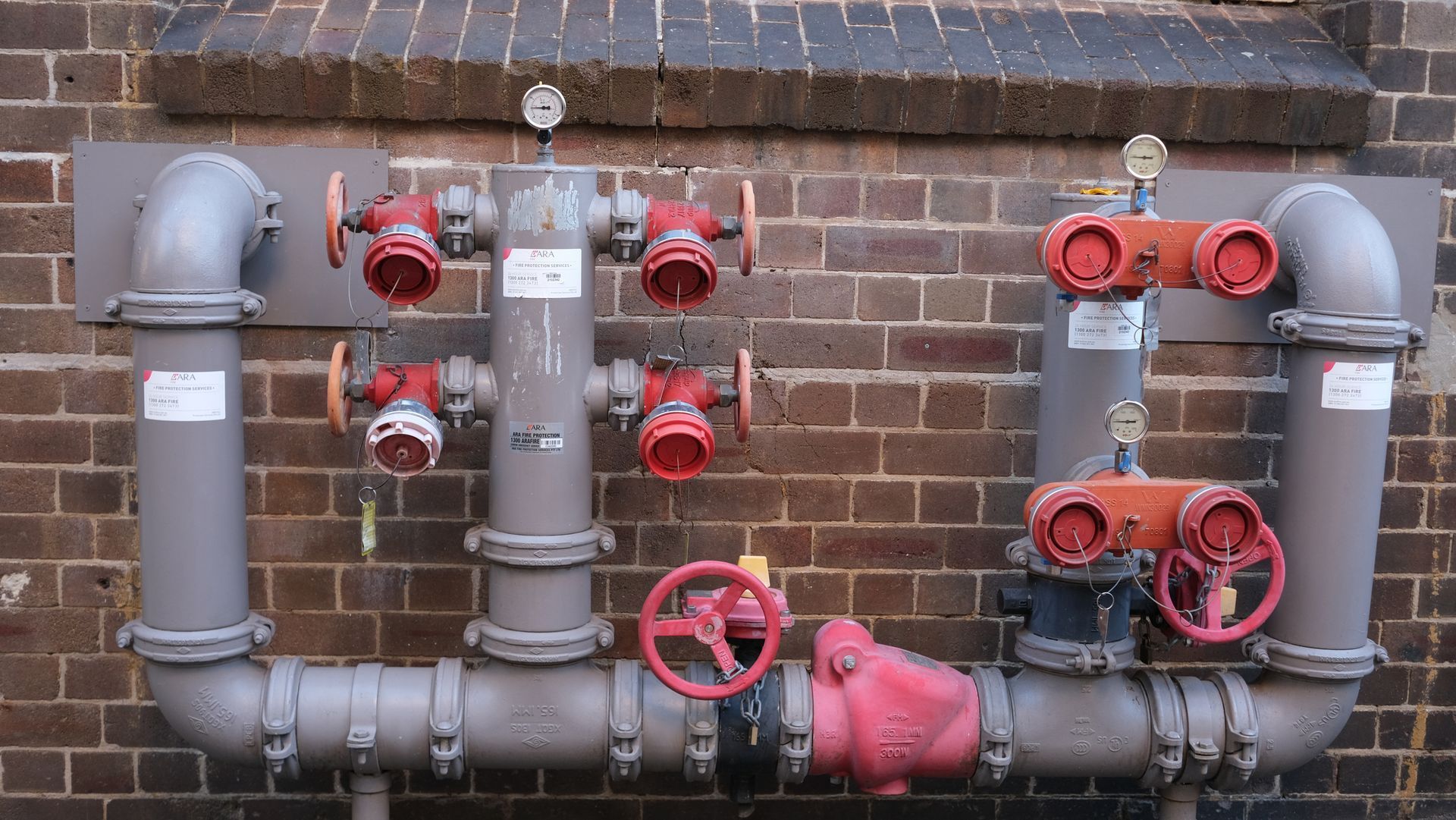 Outdoor brick wall with gray pipes, red valves, and a red pump assembly for plumbing or fire protection