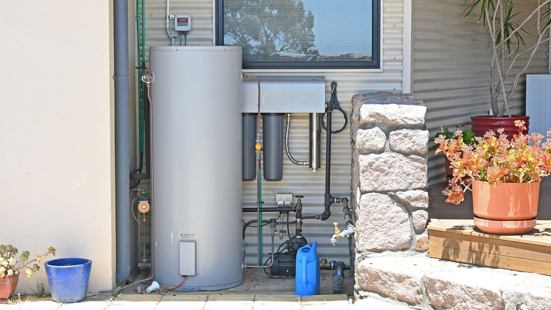 Outdoor utility area with water heater, pipes, and potted plants beside a house wall