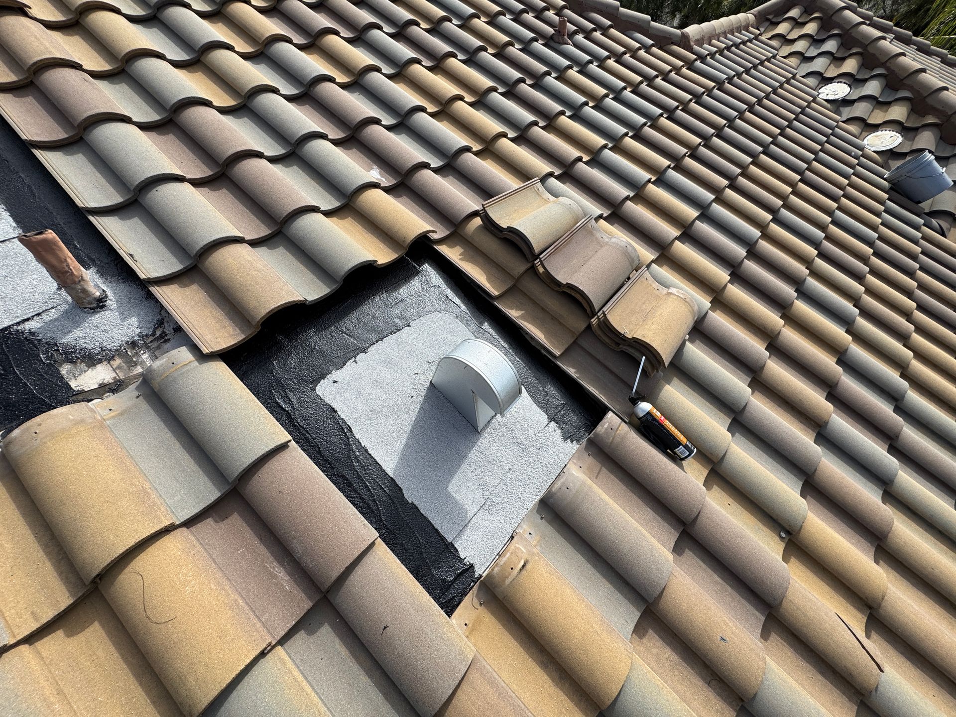 Overhead view of a clay tile roof with a square opening exposing the underlayment and a metal vent pipe. Overhead view of a clay tile roof with a square opening exposing the underlayment and a metal vent pipe.