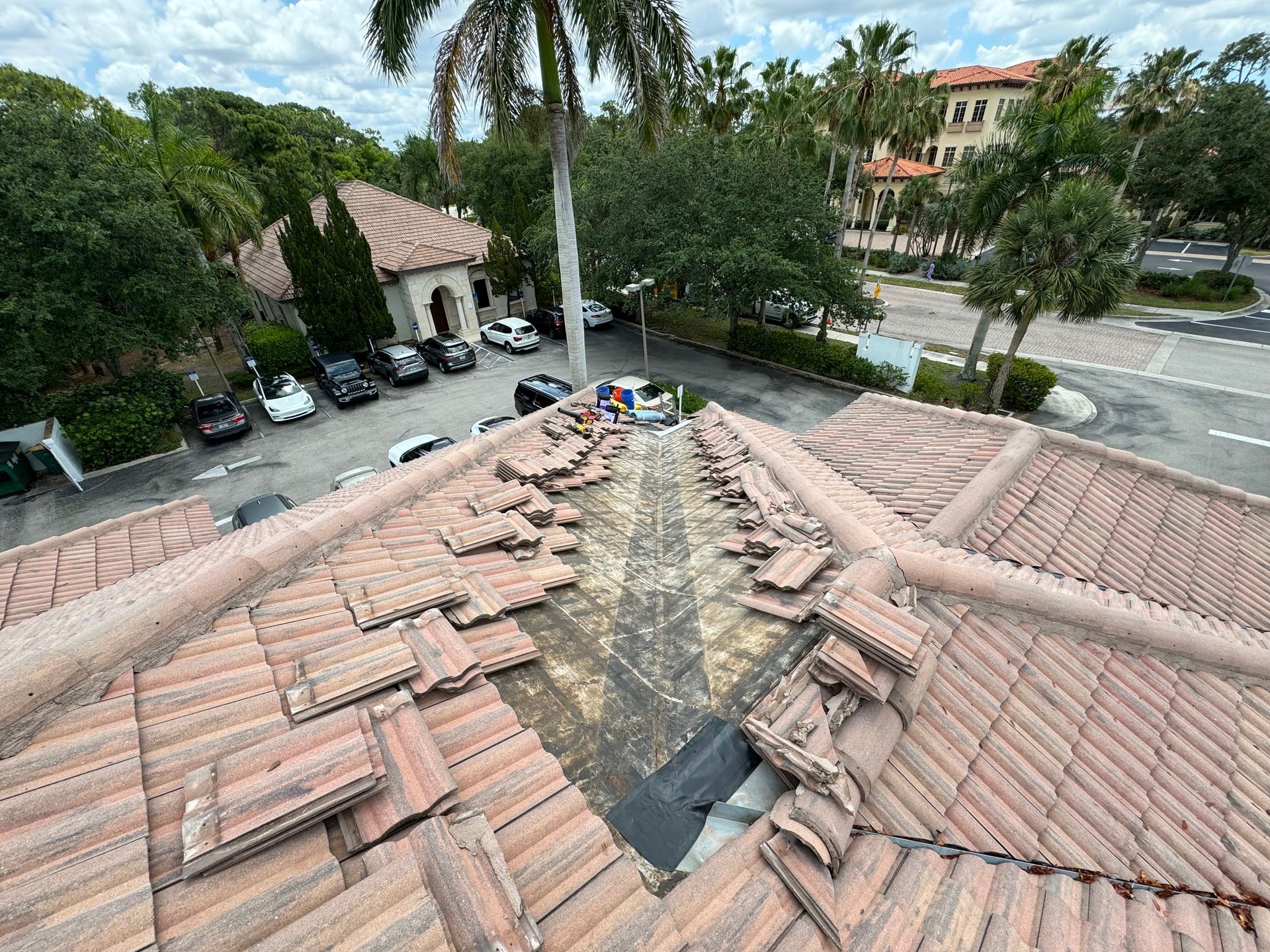 Roof tiles removed, exposing underlayment. Workers near a building with parked cars, palm trees, and blue sky. Roof tiles removed, exposing underlayment. Workers near a building with parked cars, palm trees, and blue sky.